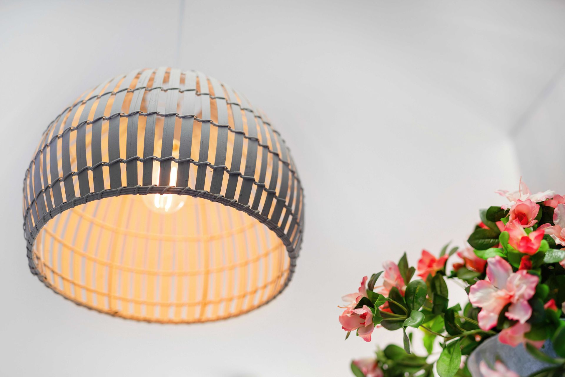 Woven dome pendant lamp with warm light beside pink flowers on a white wall