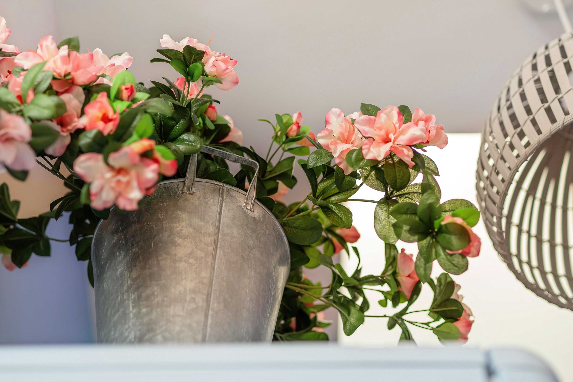 Pink flowers in a gray pot on a white shelf, with a woven lamp shade on the right.