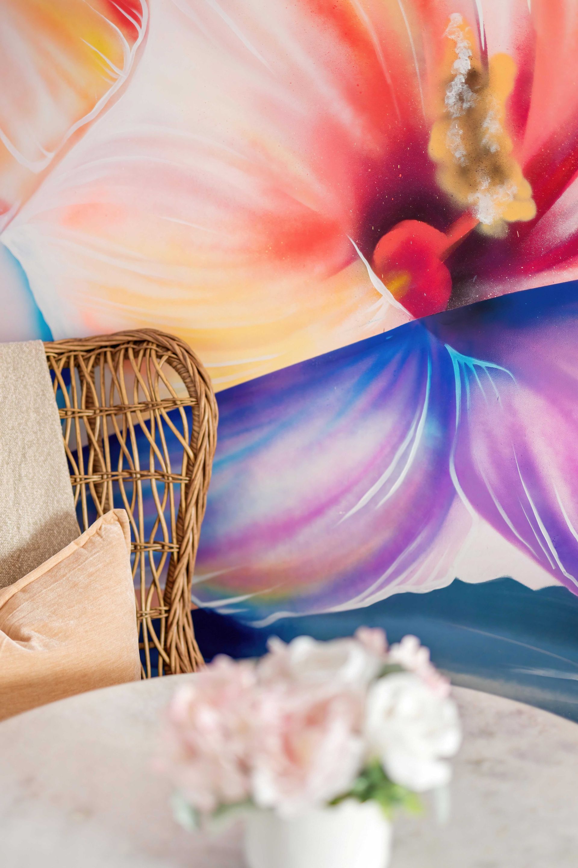 A wicker chair beside a white table with flowers in front of a colorful floral wall mural
