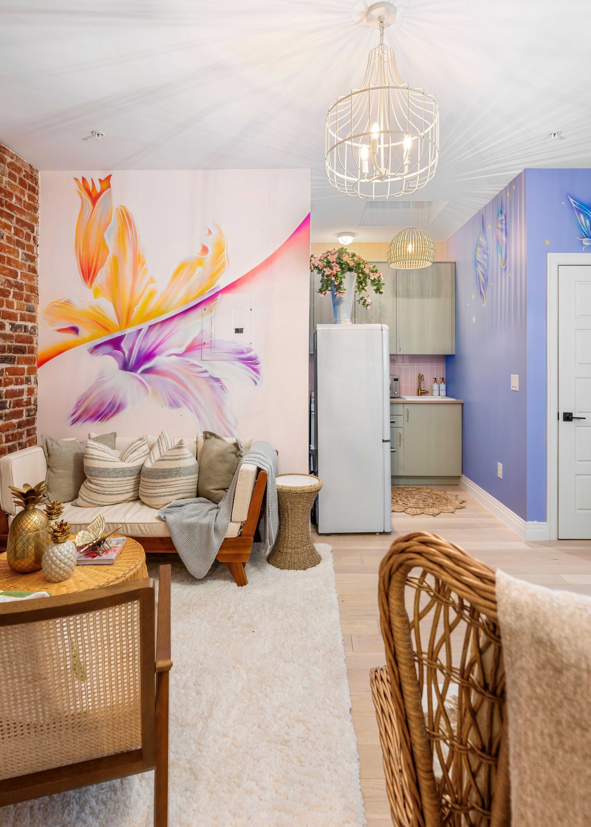 Bright boho living room with floral mural, wicker furniture, chandelier, and a small kitchen nook