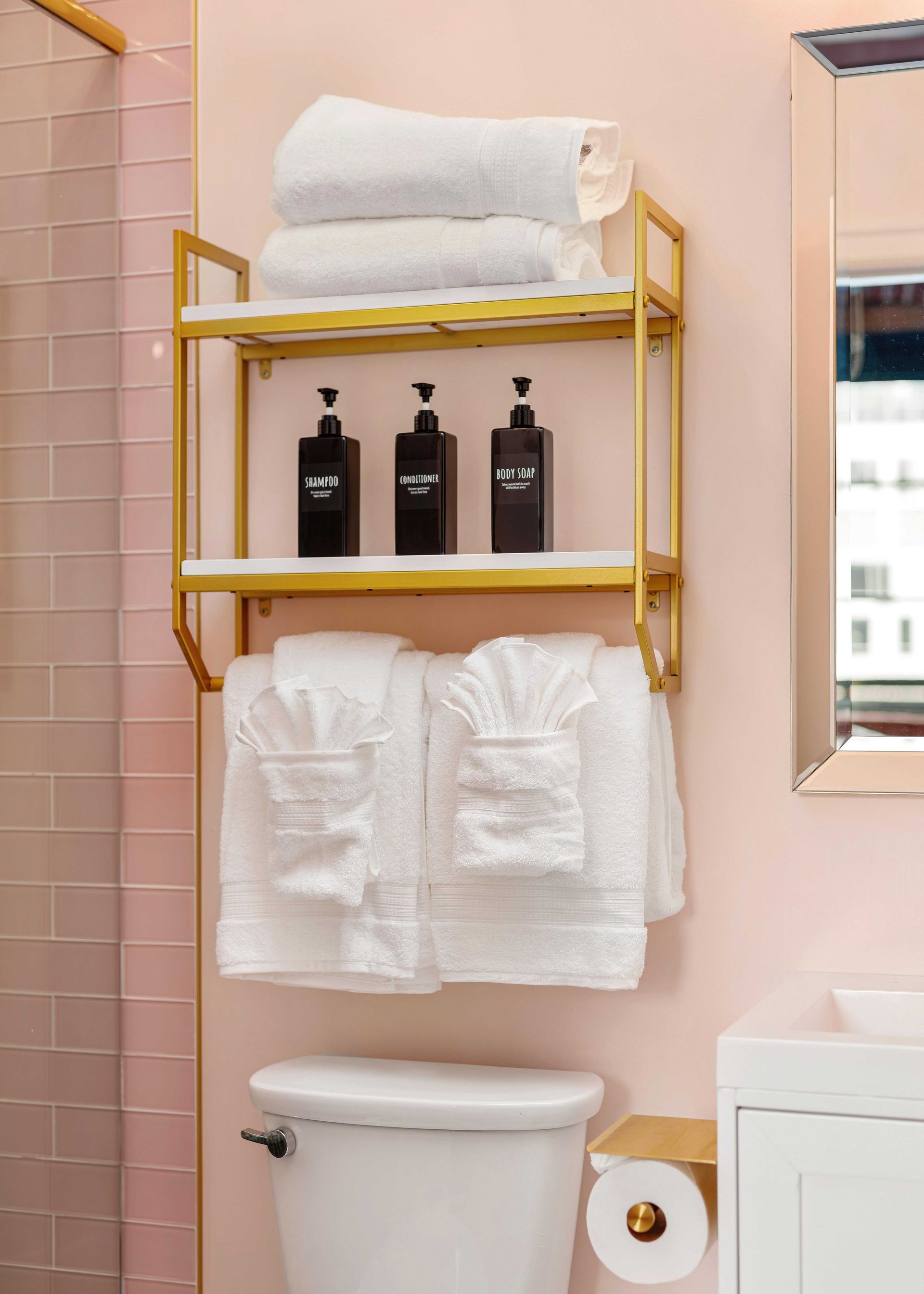 Pink bathroom with gold shelving, folded towels, black bottles, and a toilet paper roll beside a toilet