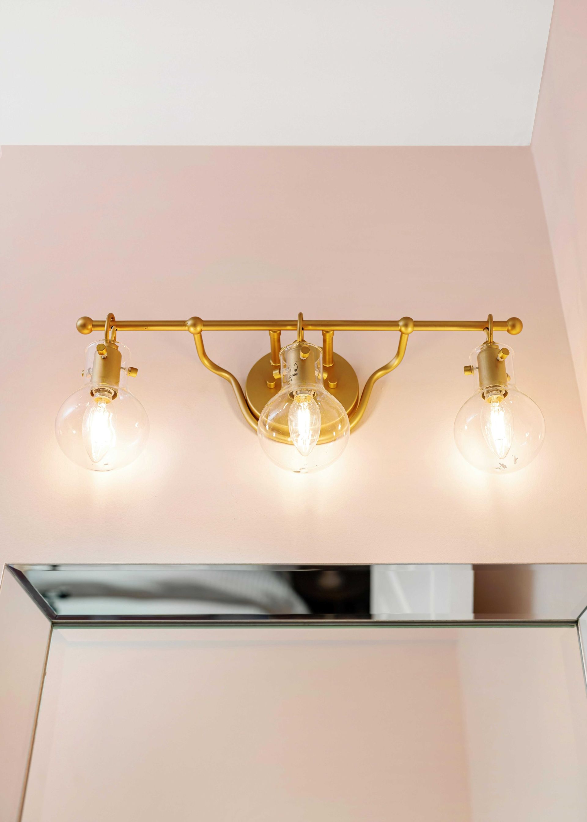 Gold vanity light with three bulbs above a bathroom mirror on a pale pink wall