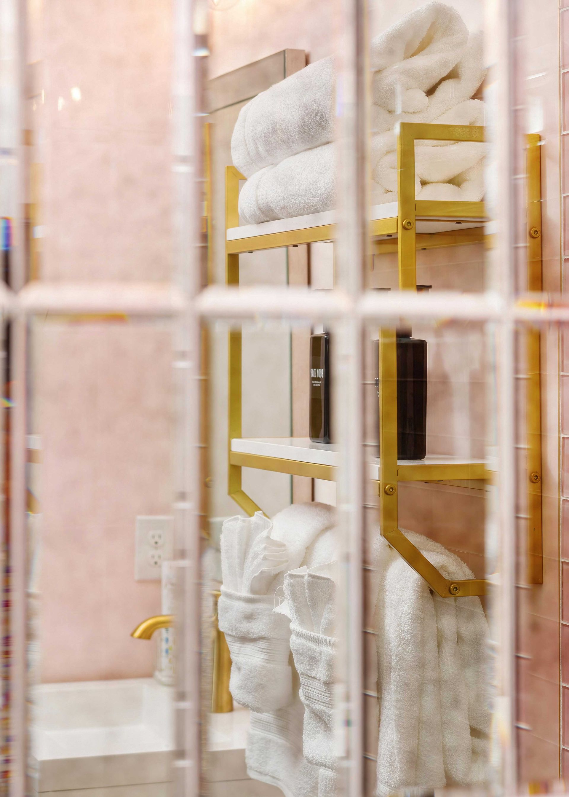 Pink bathroom with a gold towel rack and white towels seen through a glass shower door