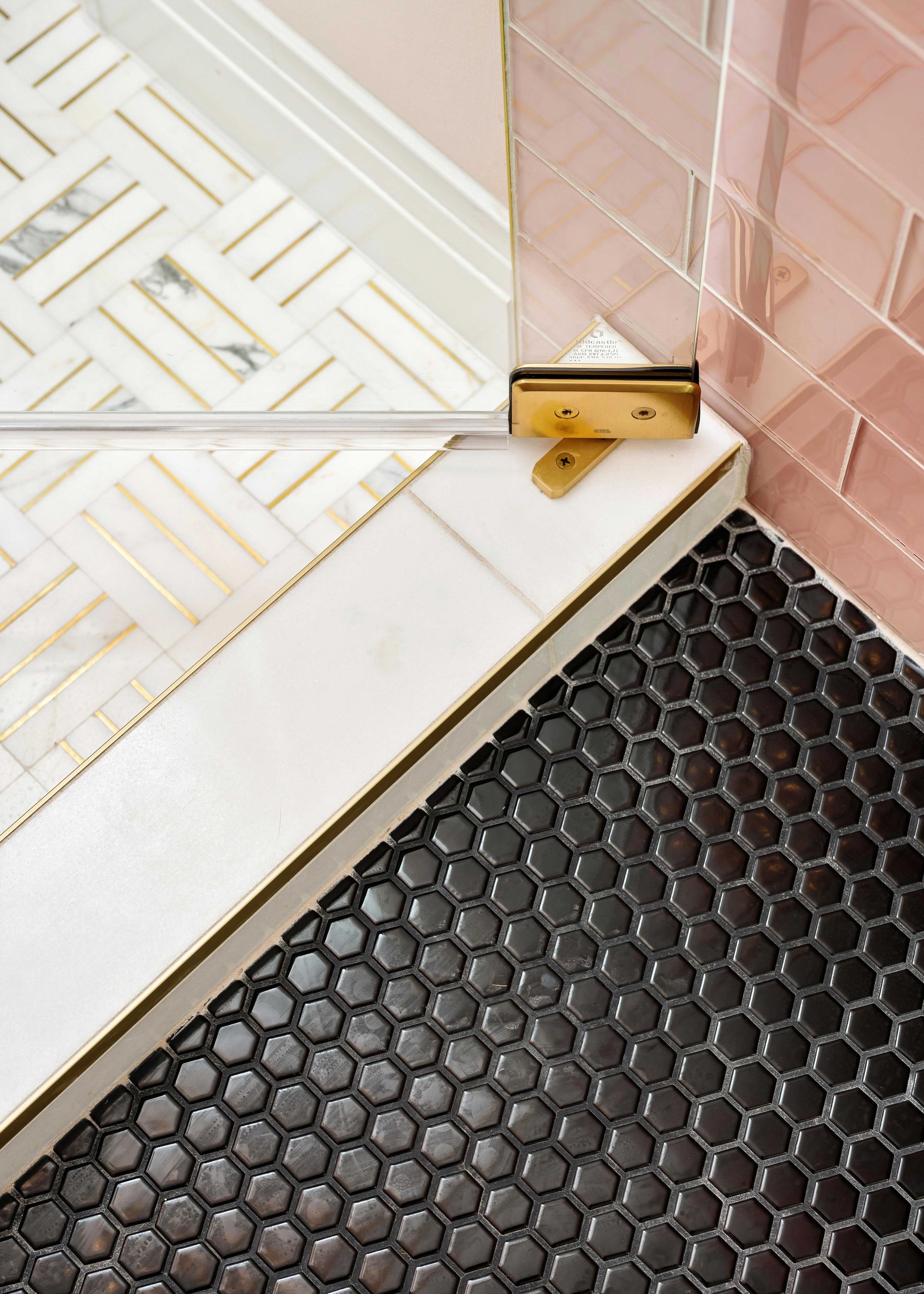 Close-up of a doorway threshold with a gold door stop, white tiled floor, pink wall tile, and black mat.