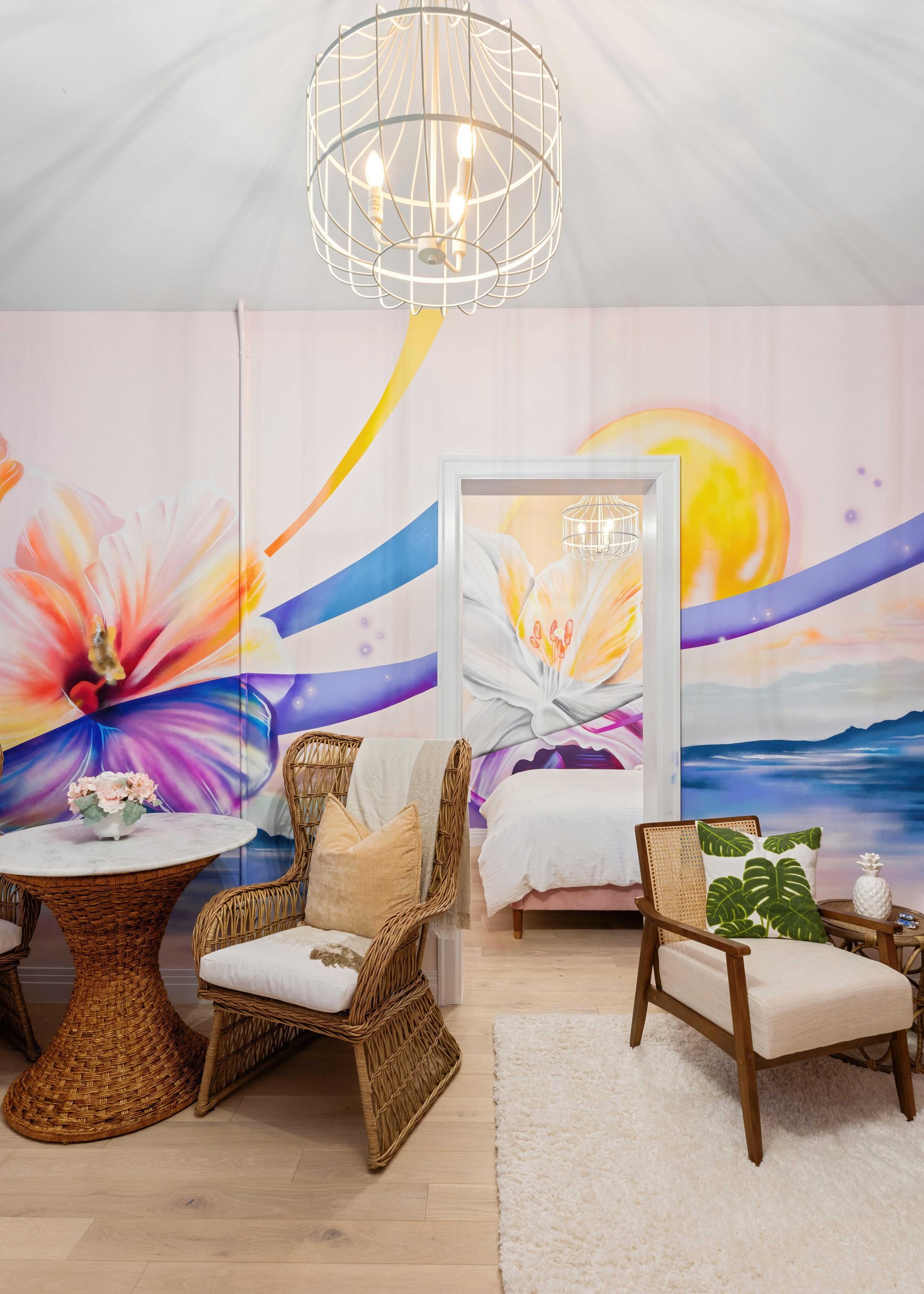 Colorful lounge setup with wicker chairs, patterned table, and abstract floral mural under a crystal chandelier