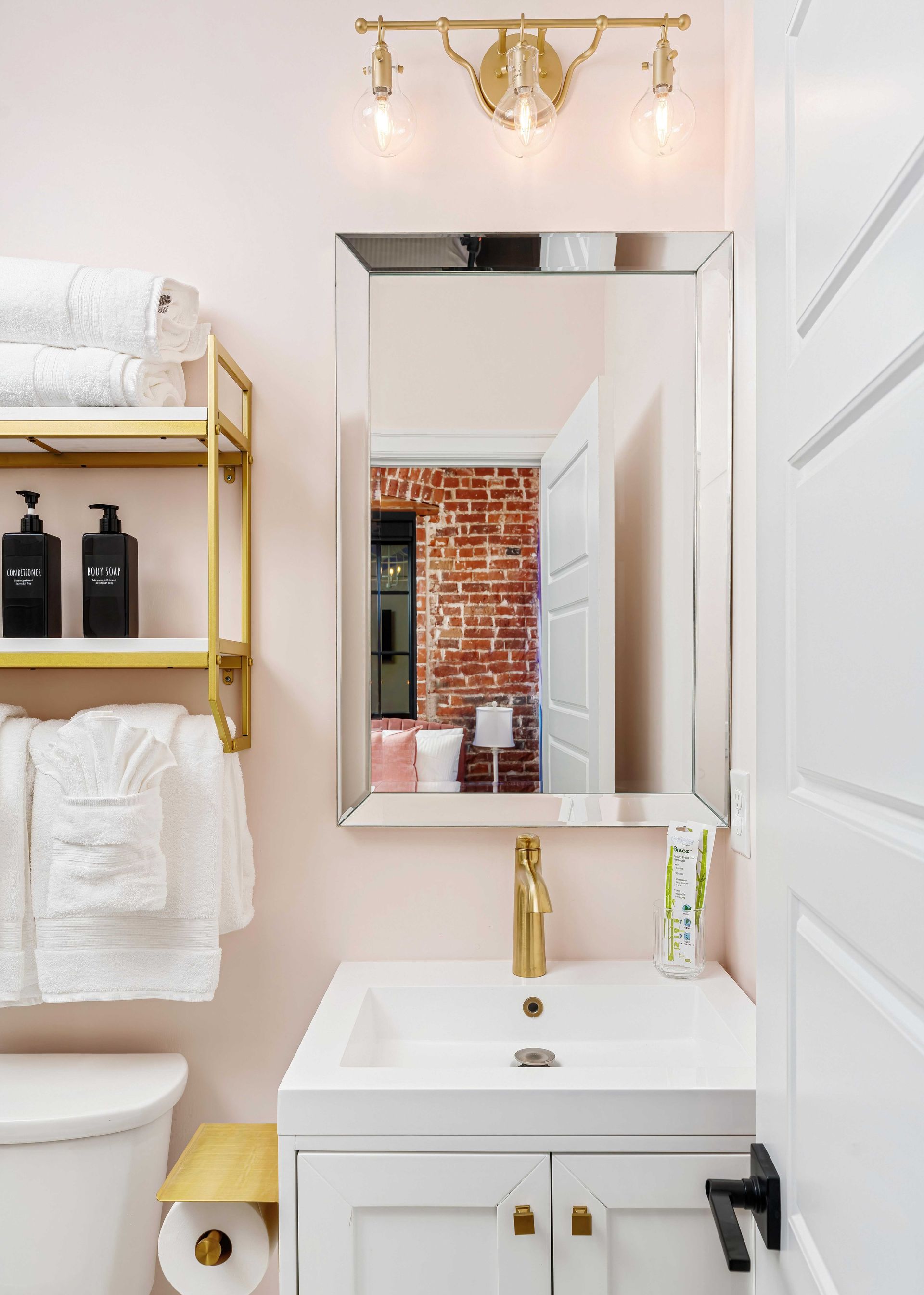Gold-accented bathroom vanity with mirror, white sink, and open shelving with towels and bottles