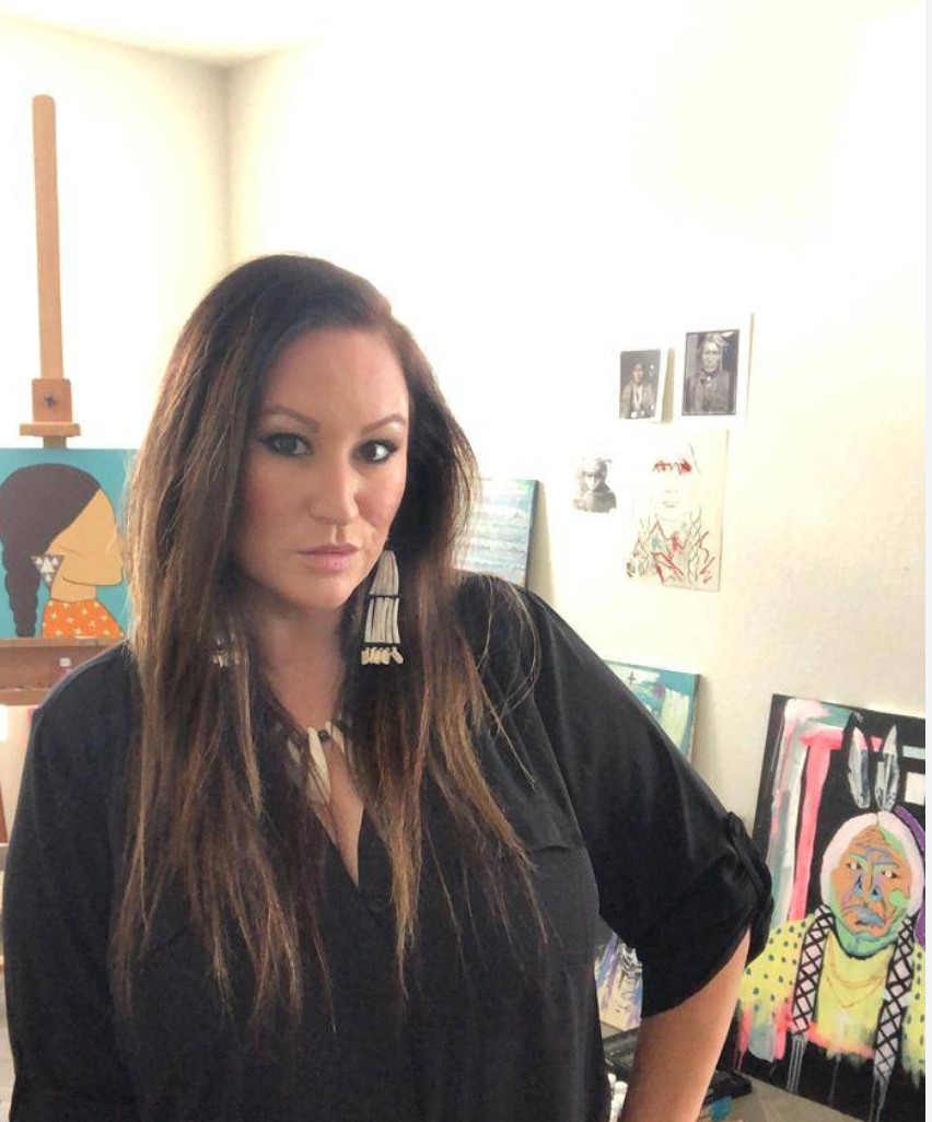 Woman standing in a bright room beside colorful artwork, wearing a black top and looking at the camera