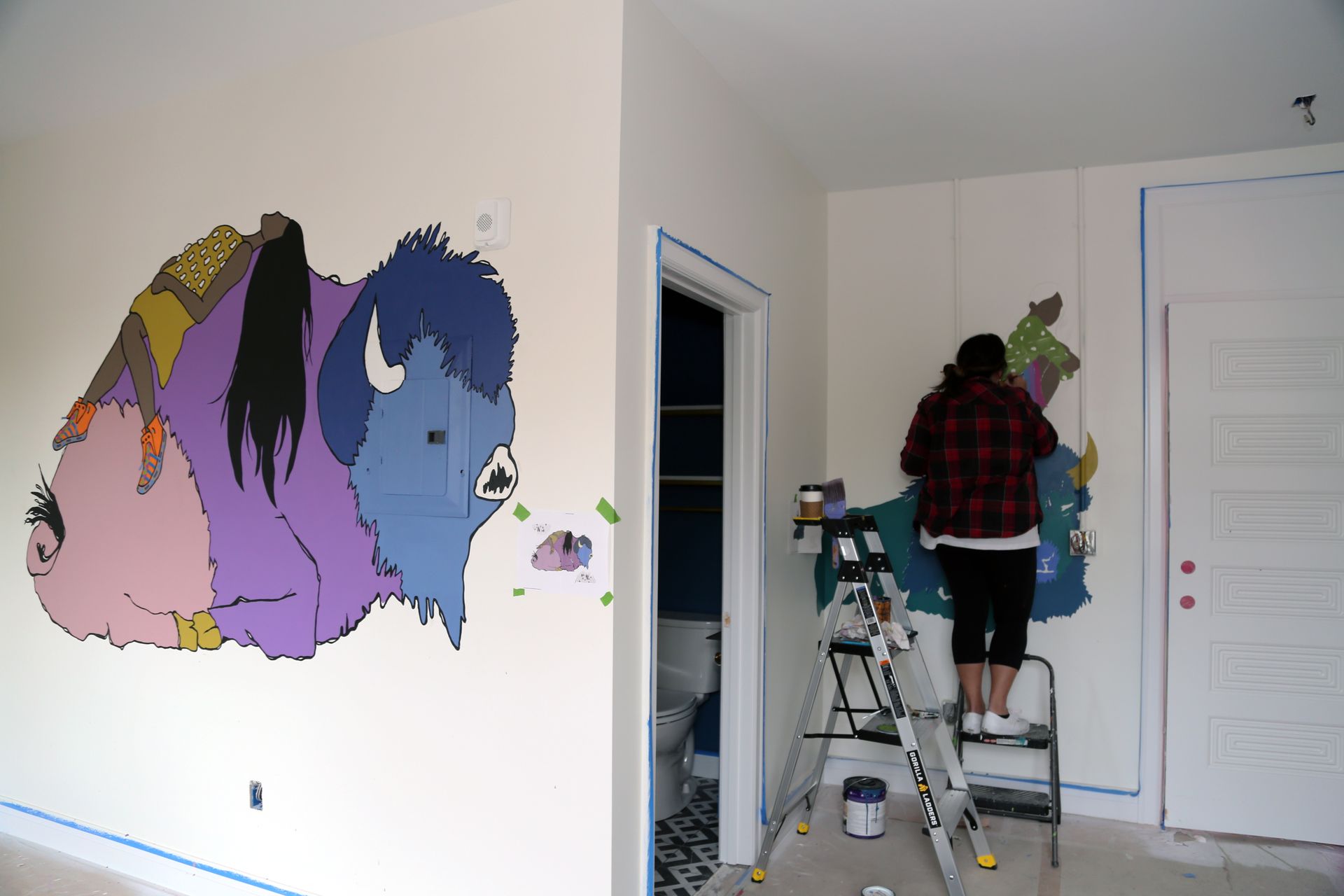 Person painting a colorful cartoon mural on a white wall beside a ladder in a room