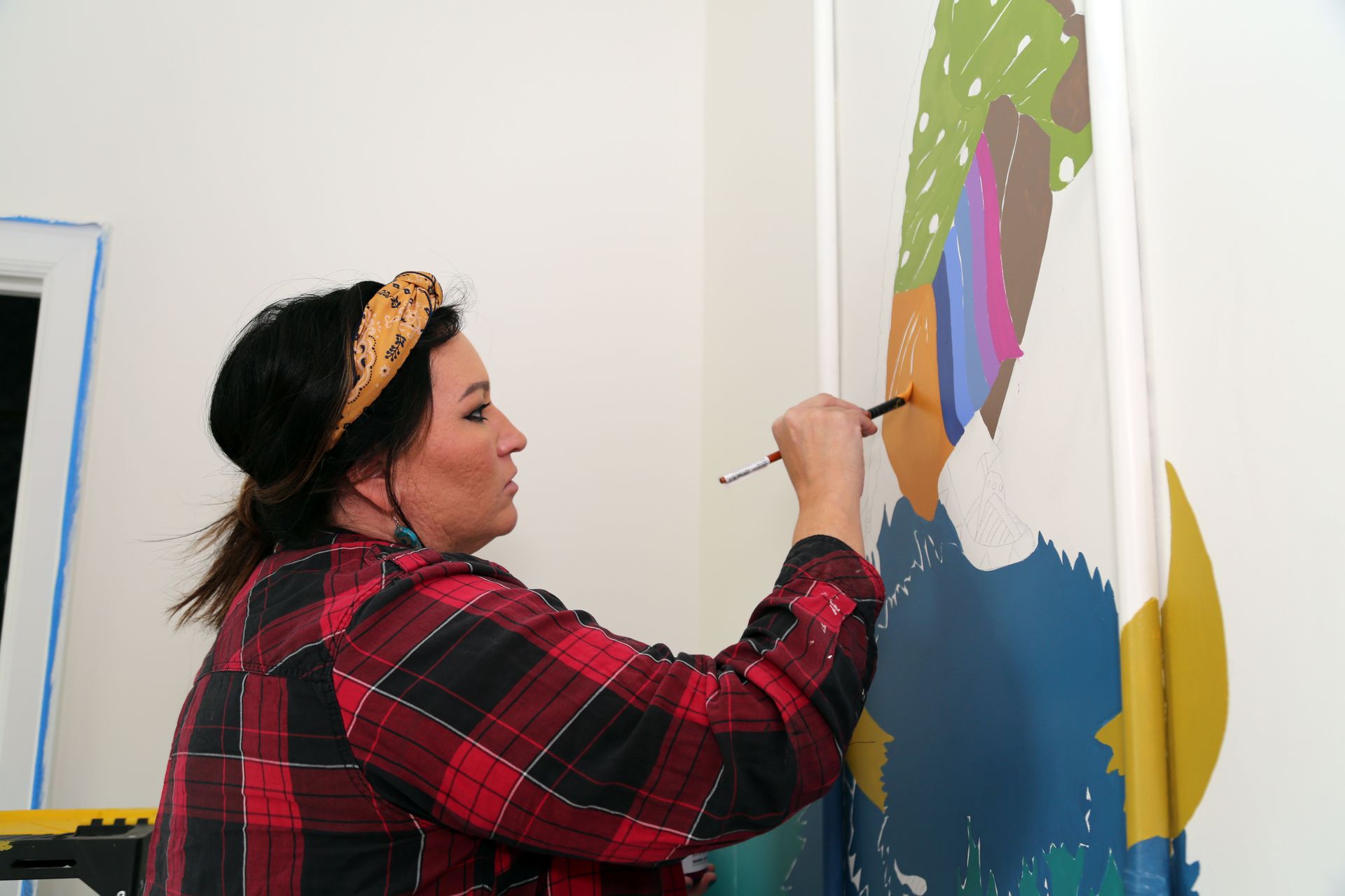 Person painting a colorful mural on a wall indoors, using a brush while wearing a red plaid shirt.