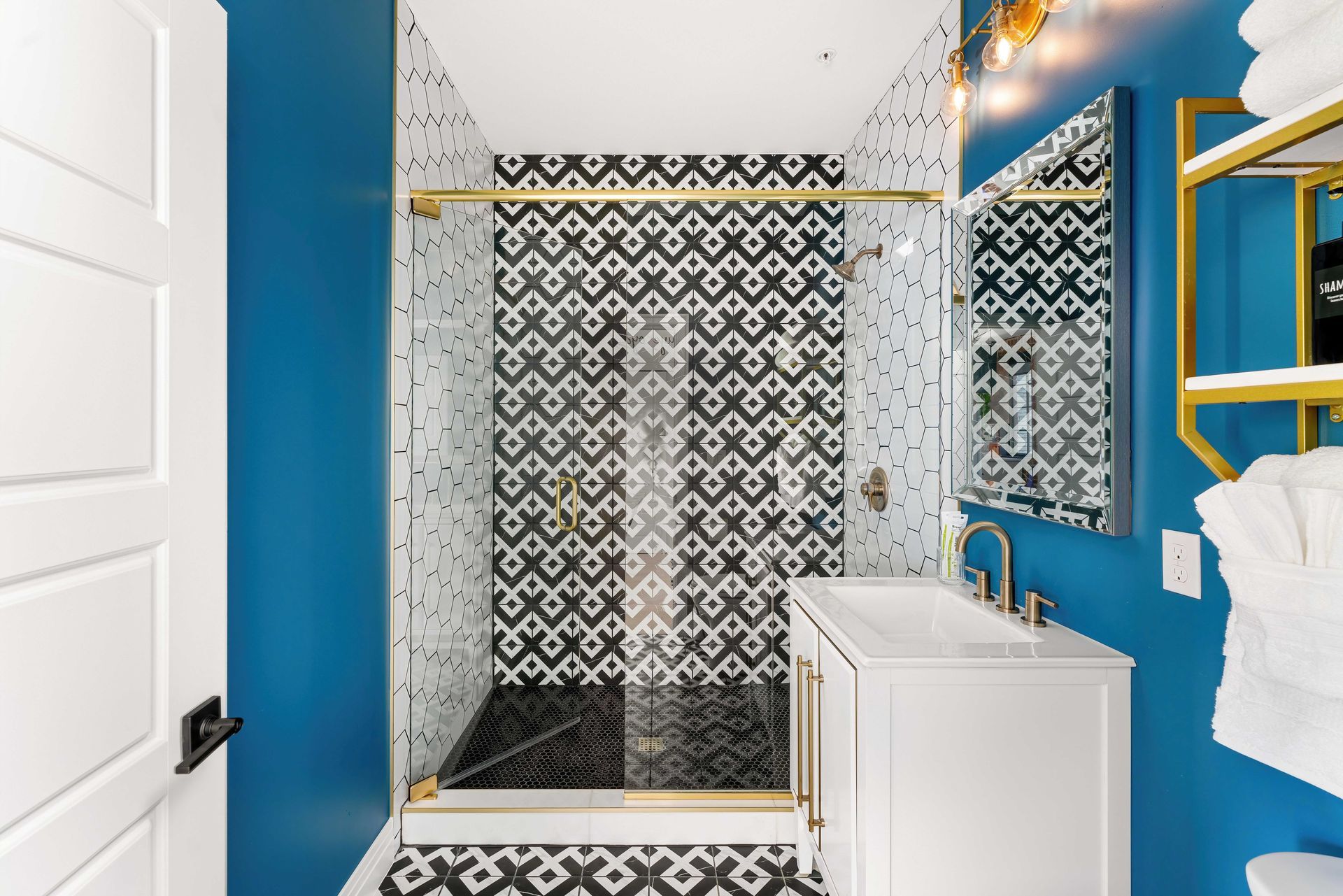 Blue bathroom with patterned black-and-white tile shower, gold accents, and a white vanity