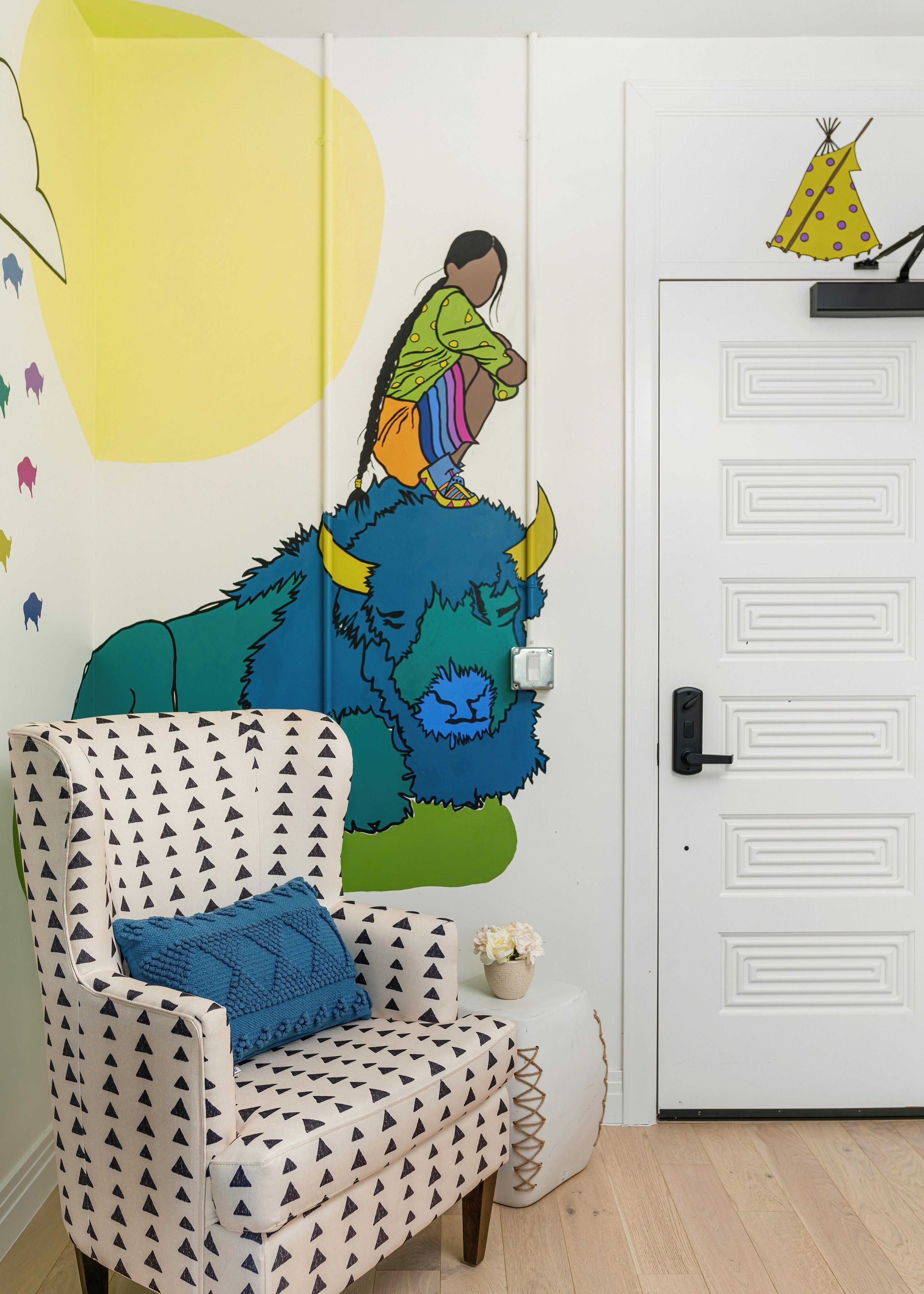 Colorful mural above a patterned armchair in a bright room with white door and wood floor