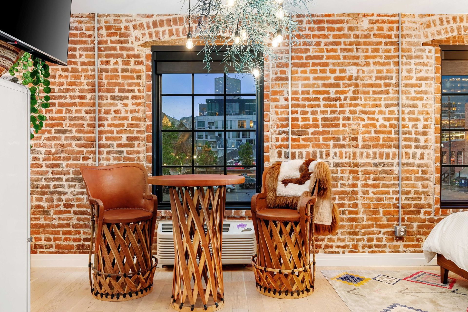 Two woven bar stools beside a small table against a brick wall with a window and rug in a bright room