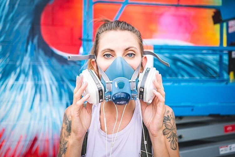 Woman wearing a respirator mask and headphones in front of a colorful graffiti wall.