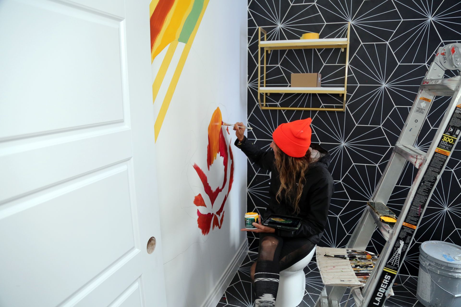 Person painting a white wall with red leaves in a small room with black geometric wallpaper.