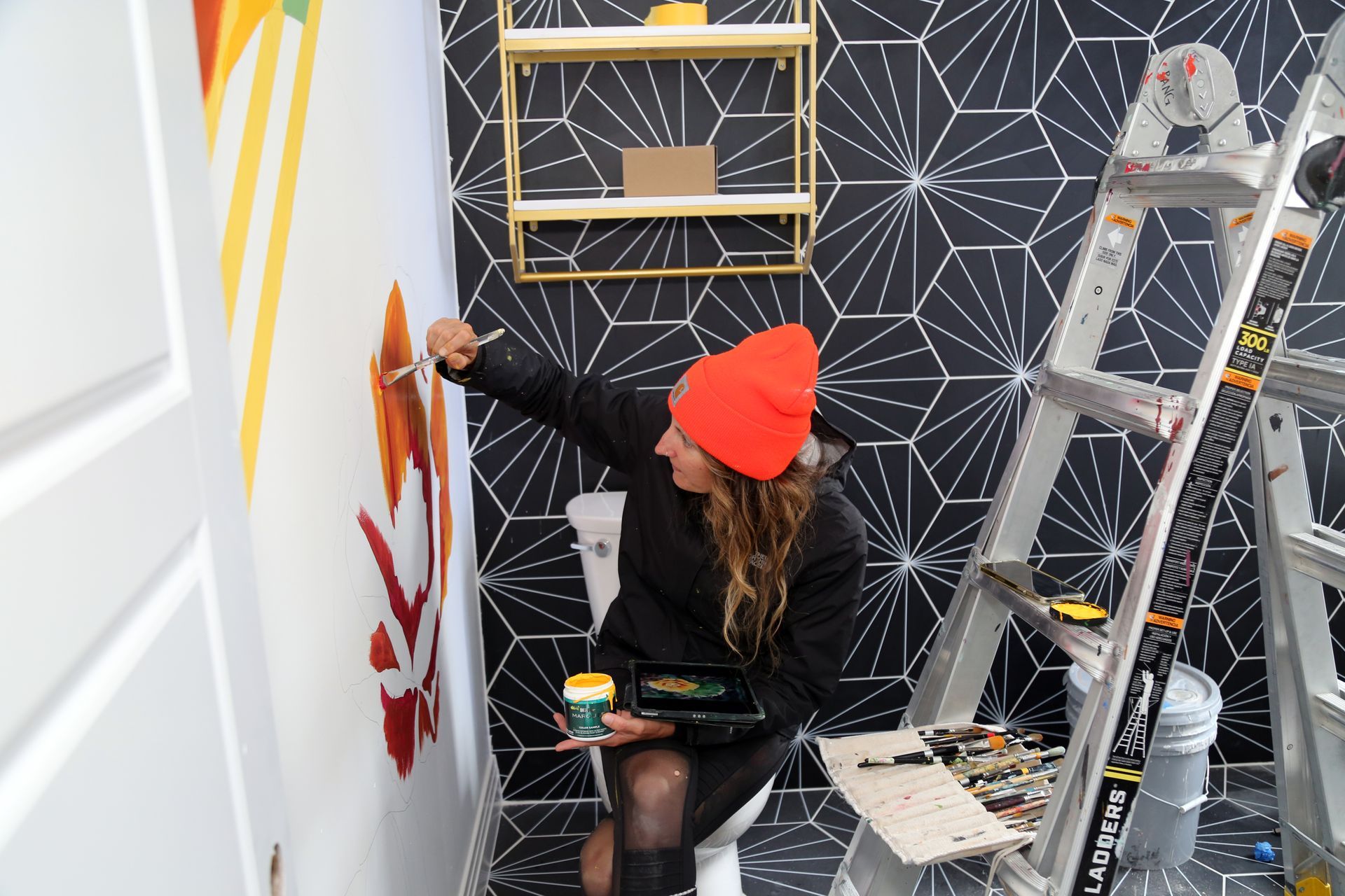 Person in a red beanie paints a white wall mural beside a ladder in a room with a geometric accent wall.