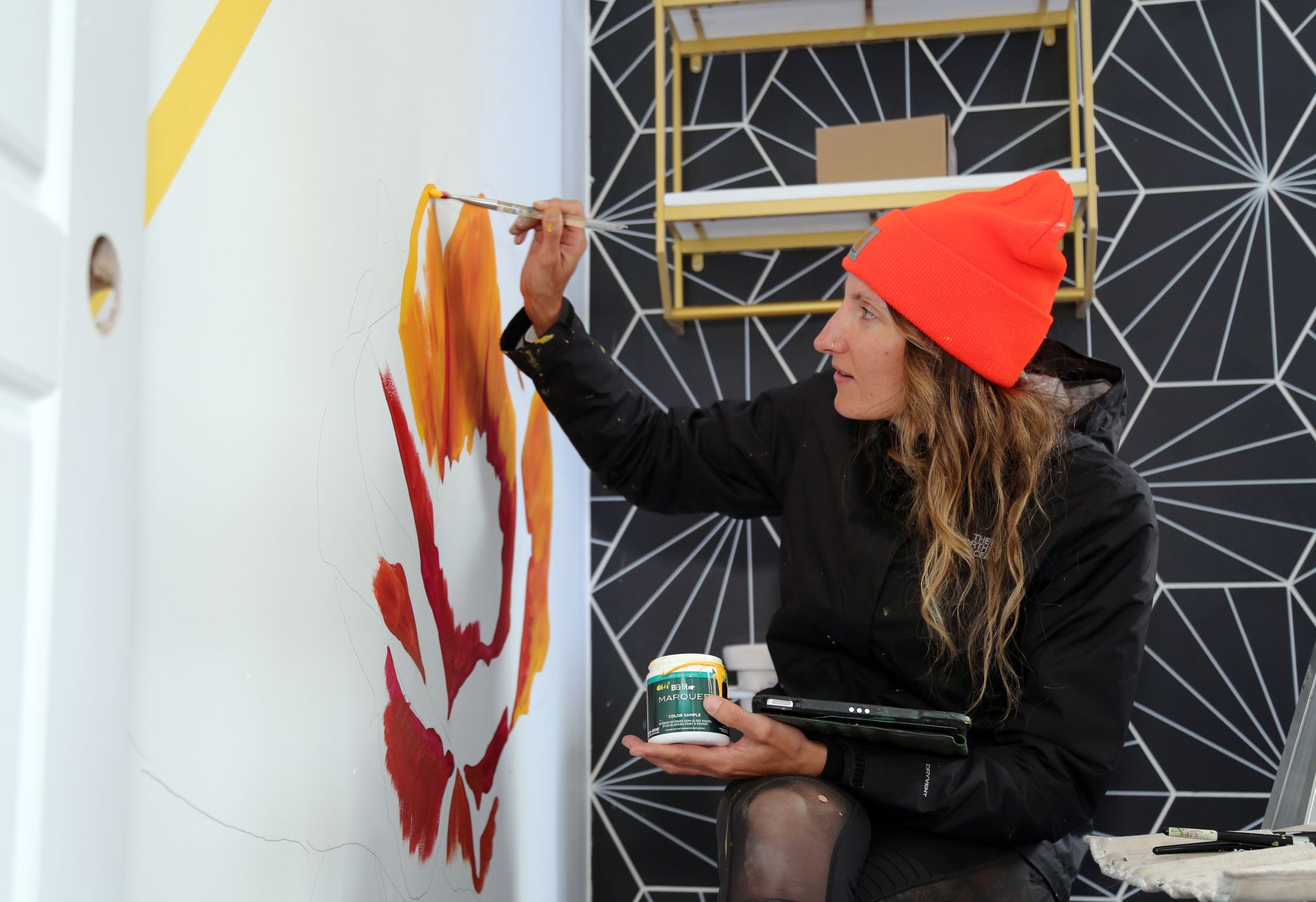 Person in orange beanie painting a mural on a black-and-white patterned wall outdoors