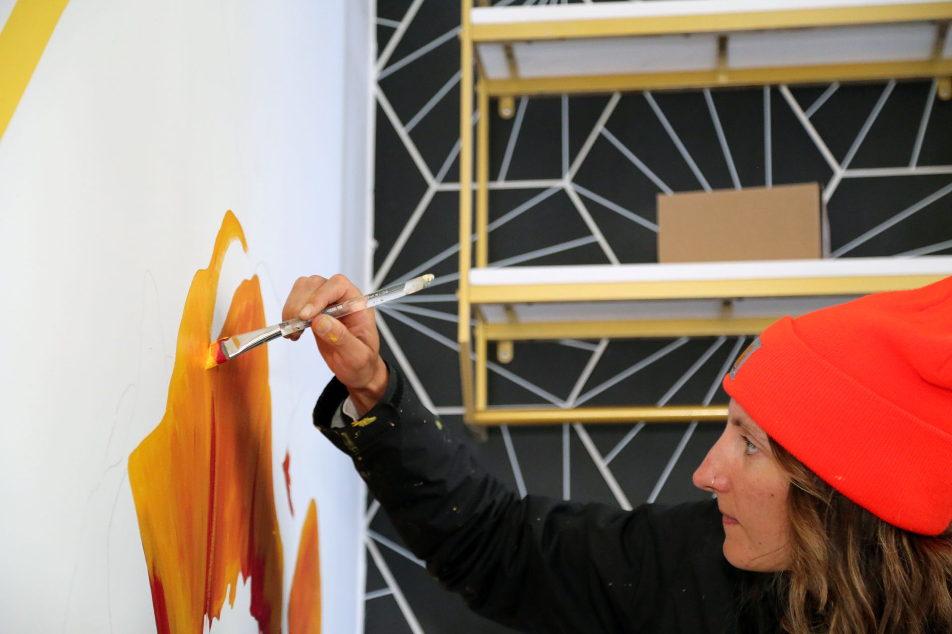 Person in a red beanie painting orange strokes on a wall with a brush