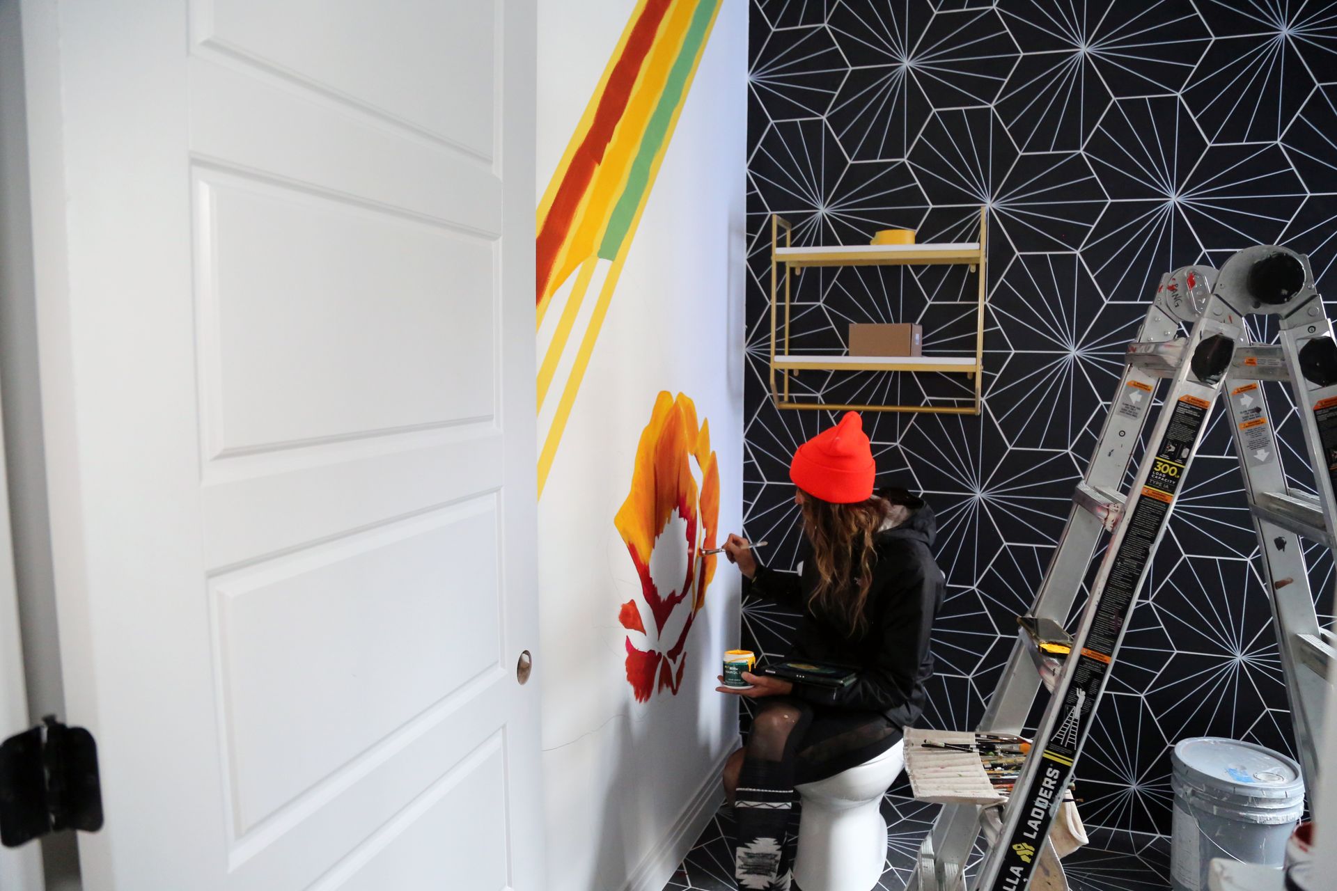 Person painting a wall mural in a room with black geometric wallpaper and a ladder nearby