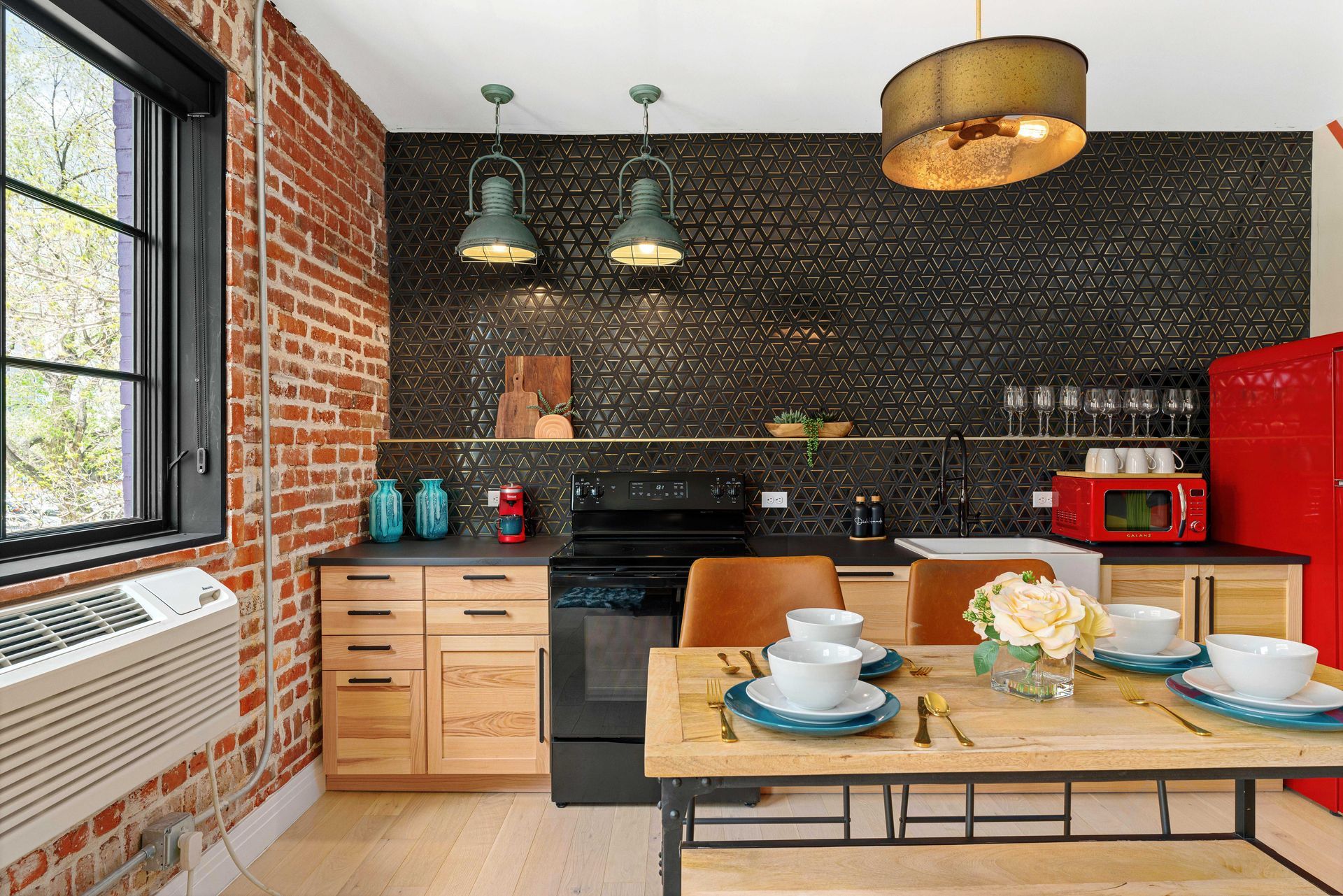Modern kitchen with brick wall, black tile backsplash, wood cabinets, red fridge, and a set dining table