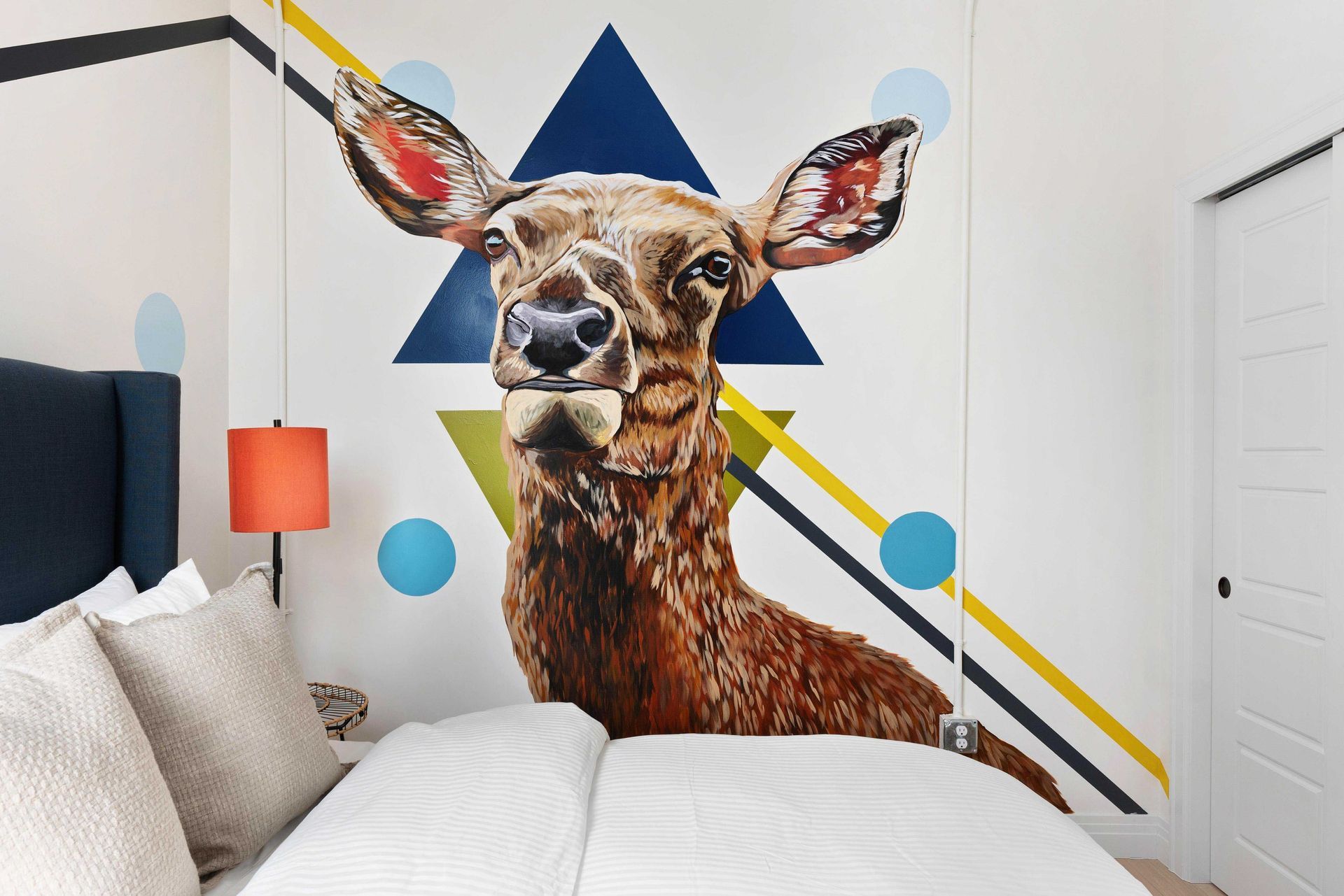 Bedroom wall mural of a deer’s face with geometric shapes, beside a bed and orange lamp
