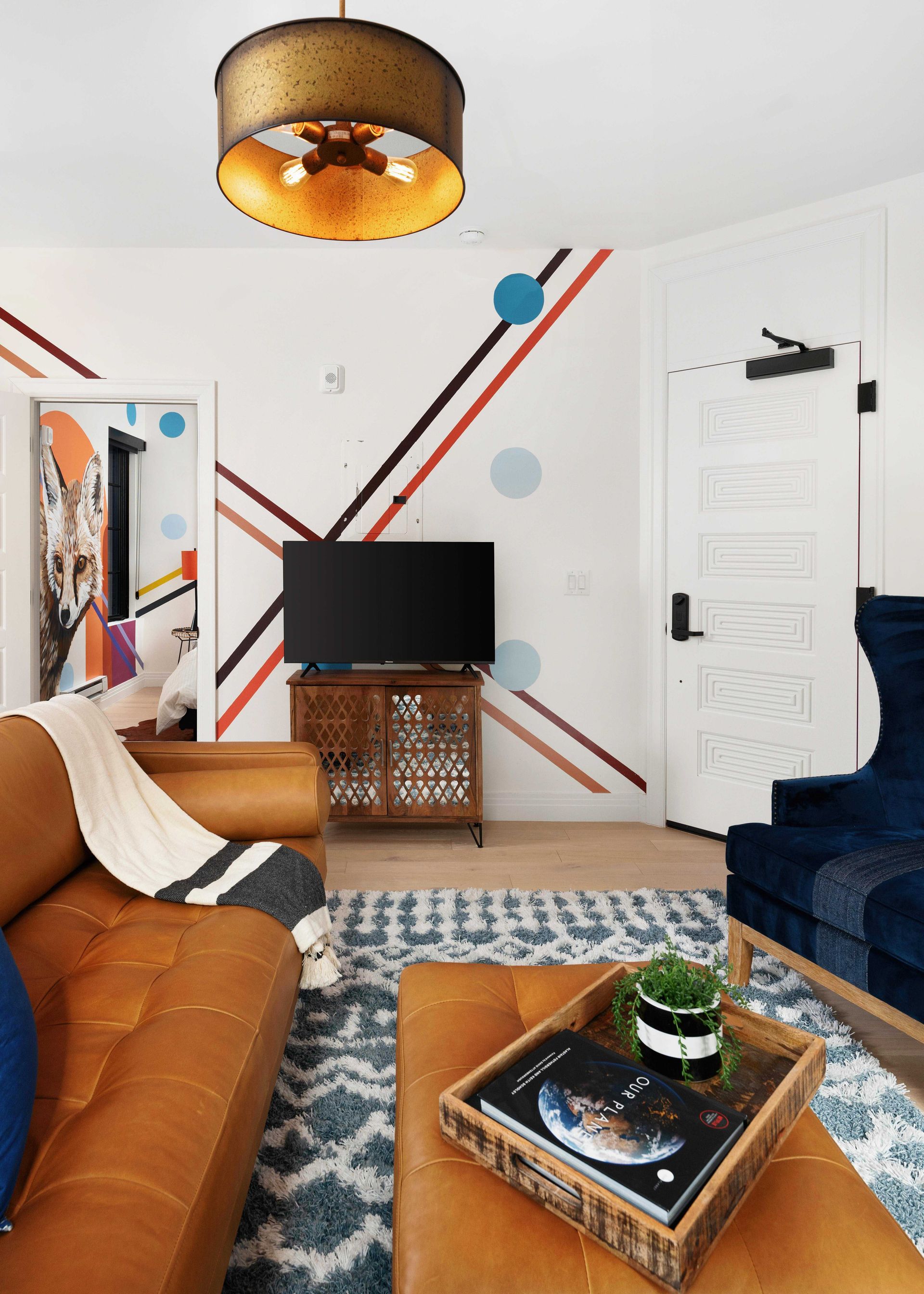 Modern living room with orange sofa, blue chair, TV, patterned rug, coffee table, and white door with diagonal wall stripes