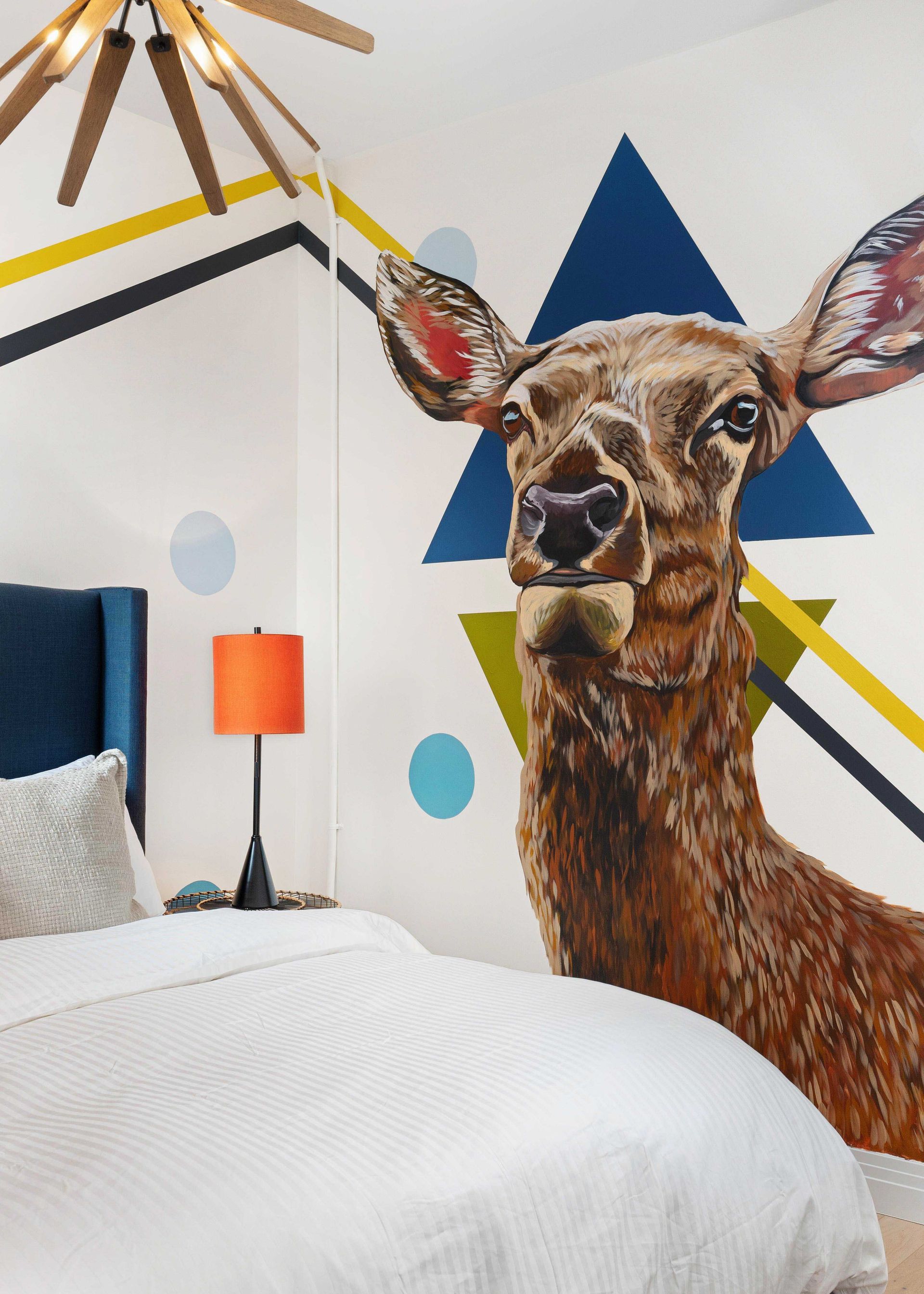 Bedroom with a large deer mural, geometric wall art, and a bed with white bedding and orange lamp.
