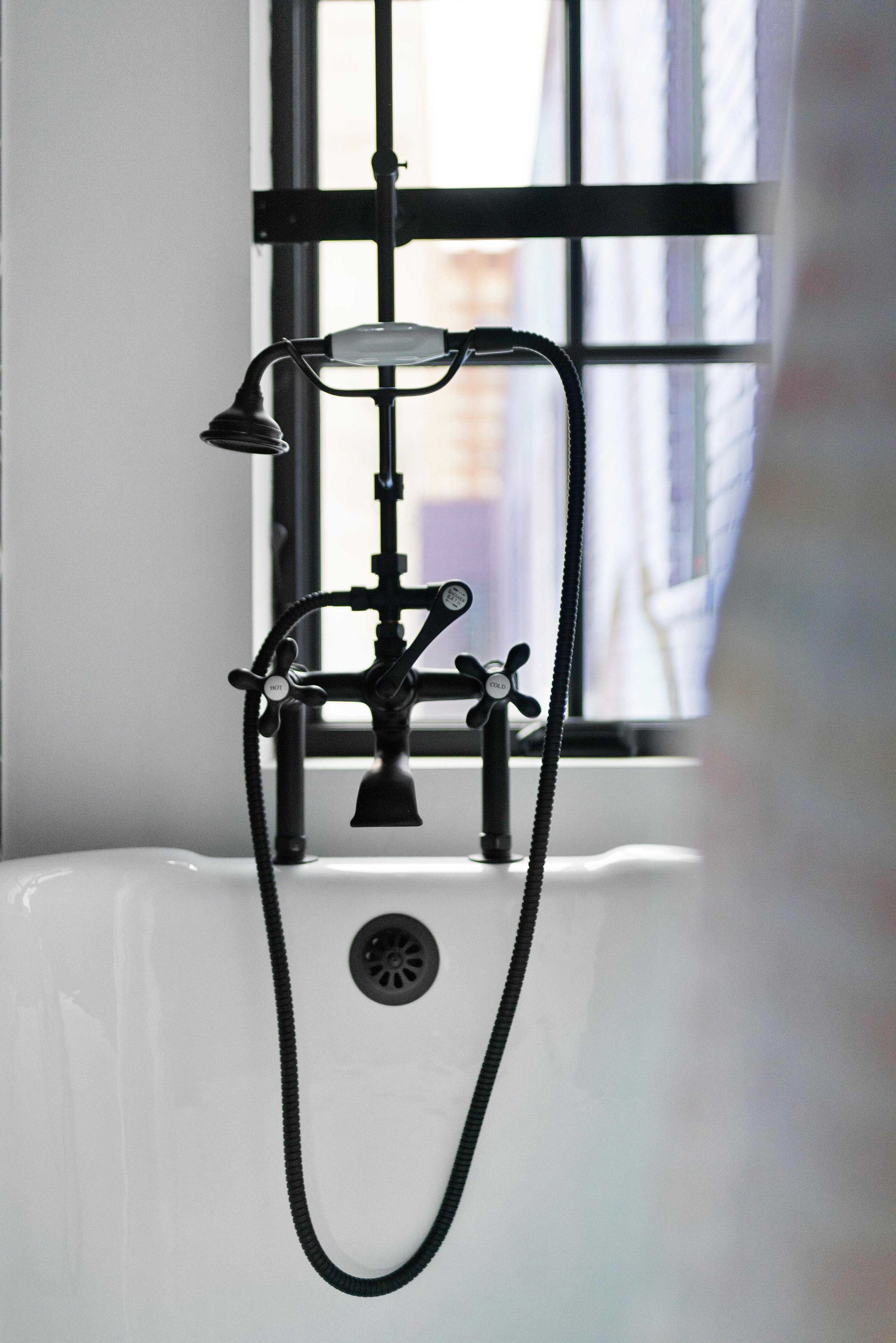 Black vintage-style bathtub faucet and shower hose in front of a window with white curtains