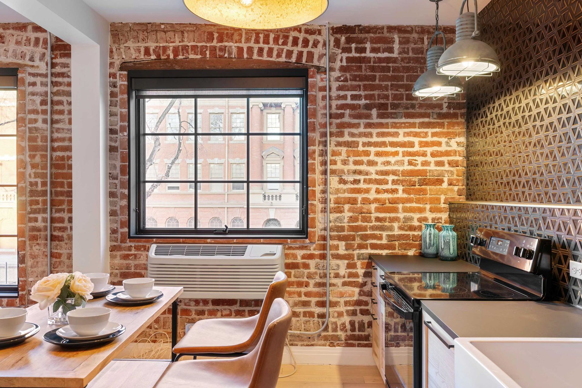 Modern kitchen with exposed brick walls, black-framed window, dining table, and pendant lights