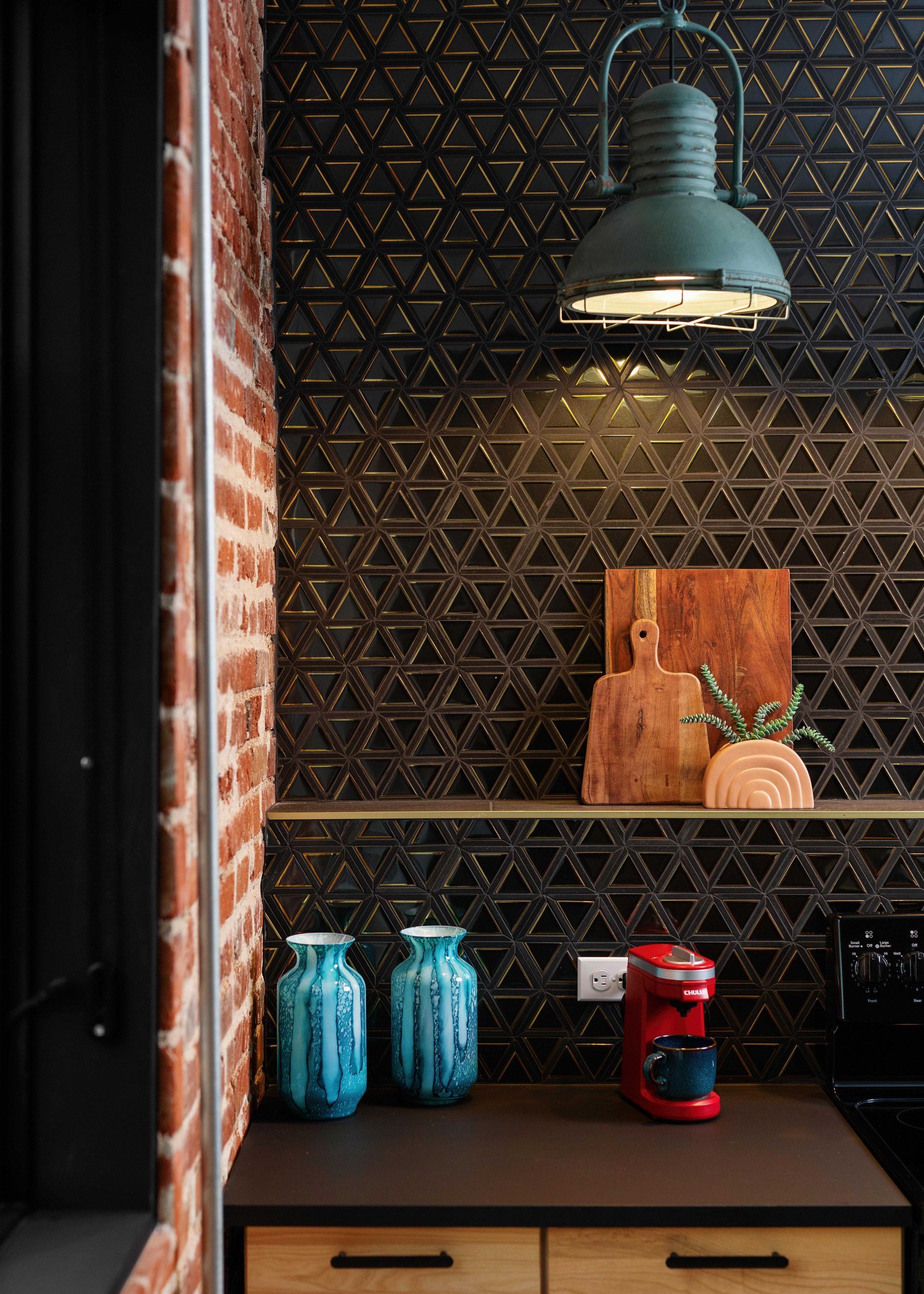 Small kitchen nook with teal pendant light, black patterned backsplash, red espresso machine, and blue jars.