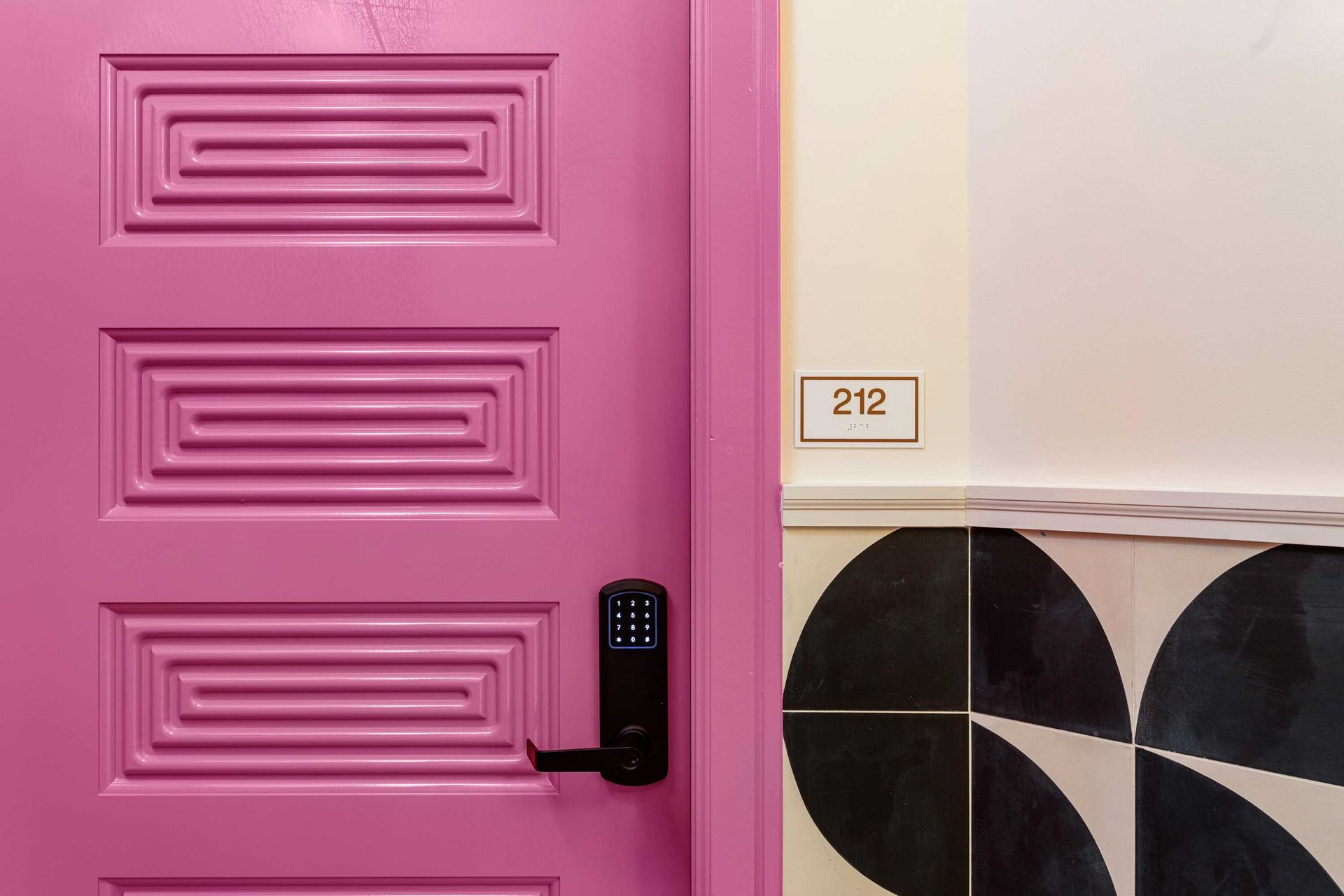 Pink front door with black handle beside a wall of black-and-white tiles and a small number plaque