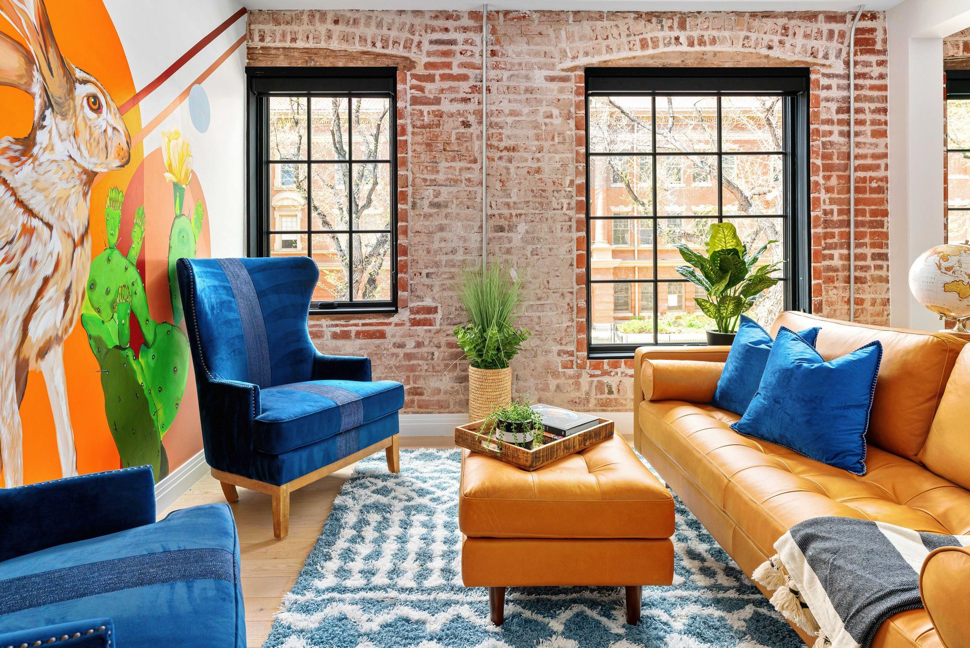 Bright living room with brick walls, blue chair, tan sofa, colorful mural, and patterned rug