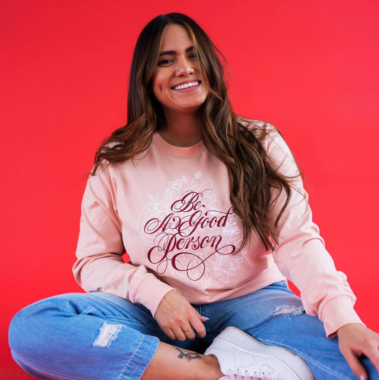 Smiling woman sitting cross-legged on a red background in a pink sweatshirt and blue jeans