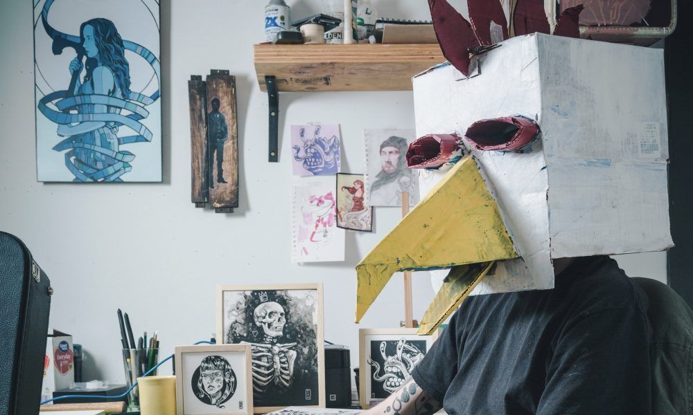 Person wearing a white box head with red eyes sits in a room with framed art on the wall.