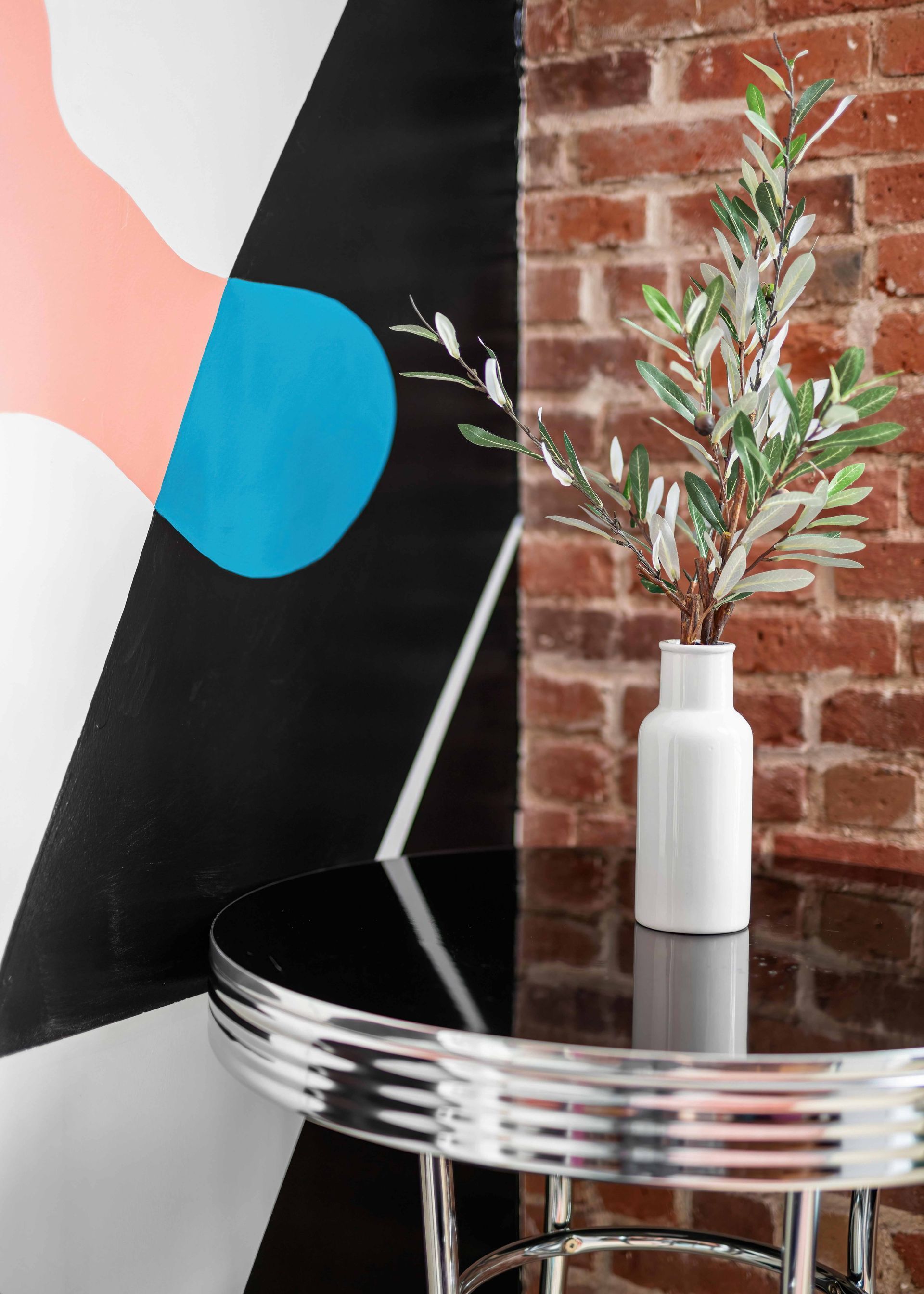 White vase with greenery on a chrome table beside a brick wall and abstract mural