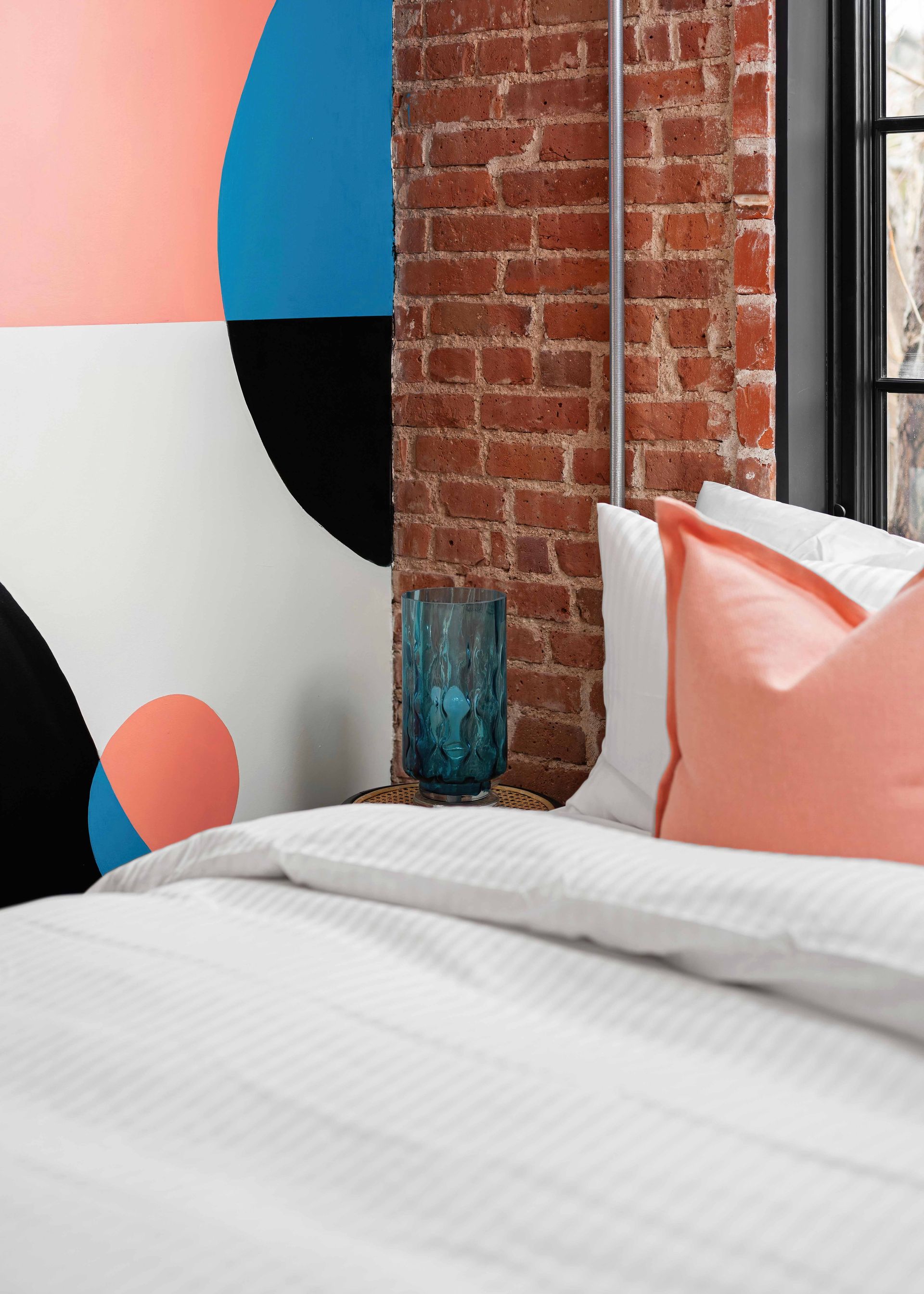 Modern bedroom with white bed, coral pillows, exposed brick wall, and colorful abstract wall art