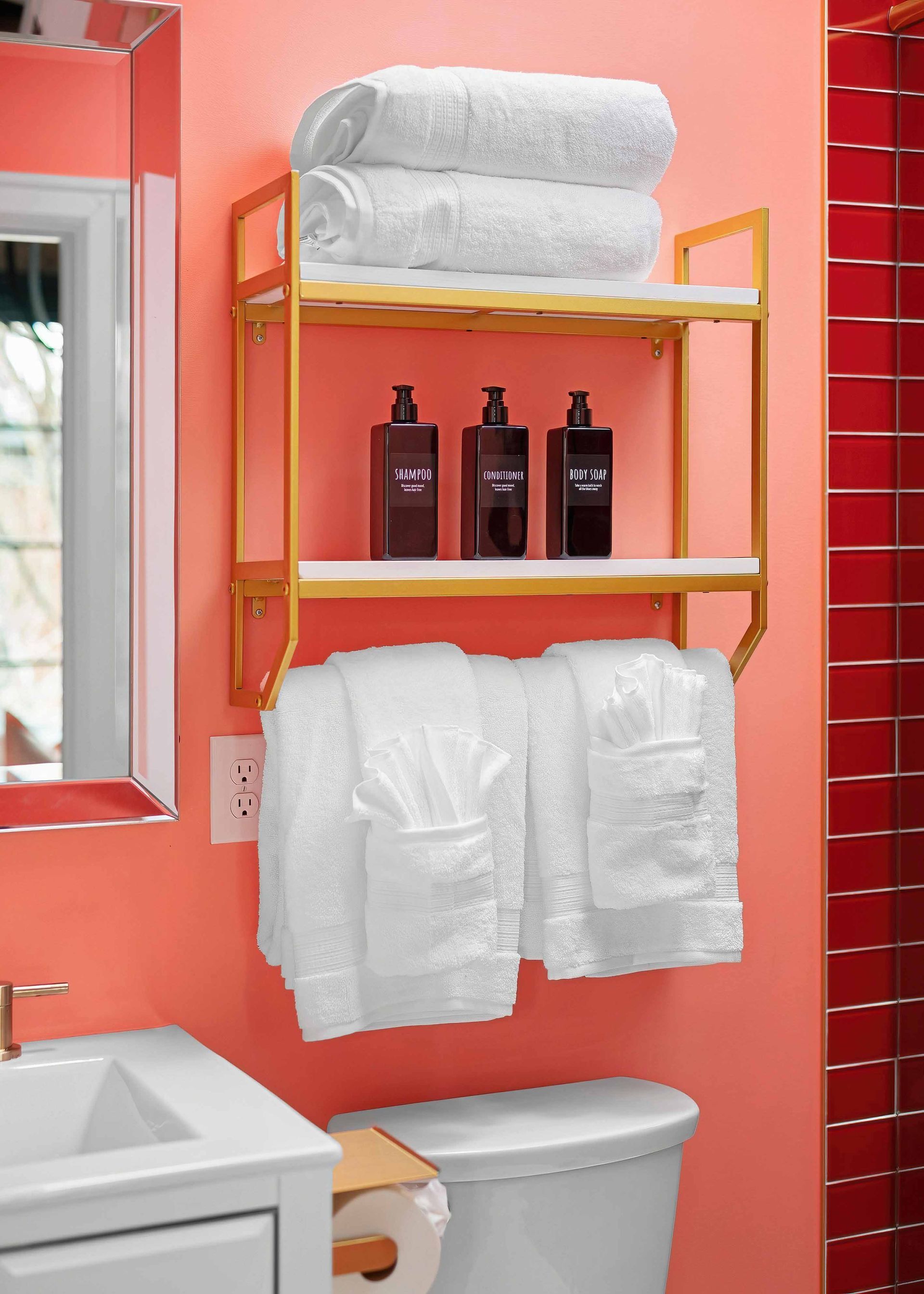 Pink bathroom wall with gold shelves holding folded white towels and toiletries above a toilet.