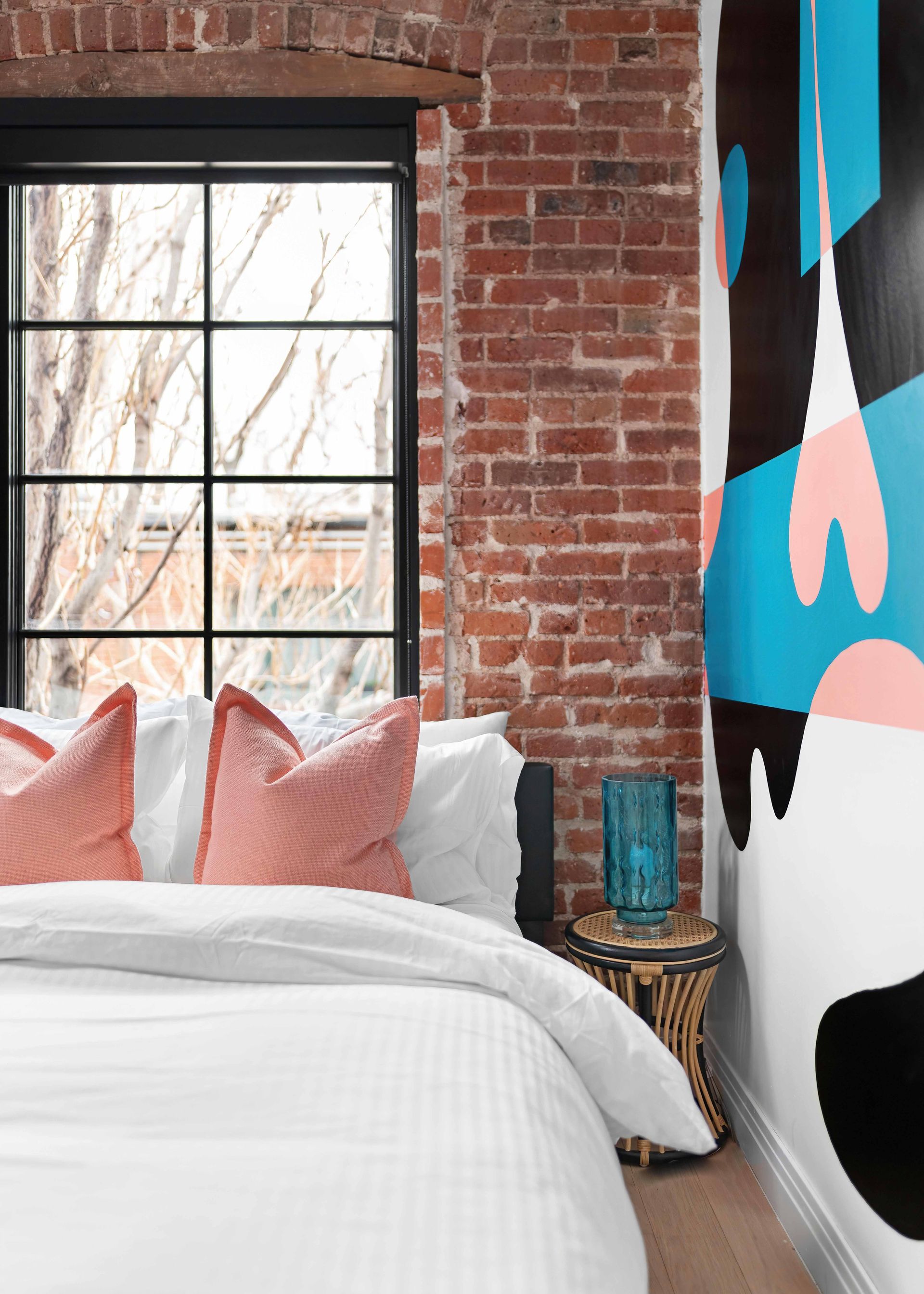 Cozy bedroom with brick wall, large window, white bedding, pink pillows, and colorful abstract wall art
