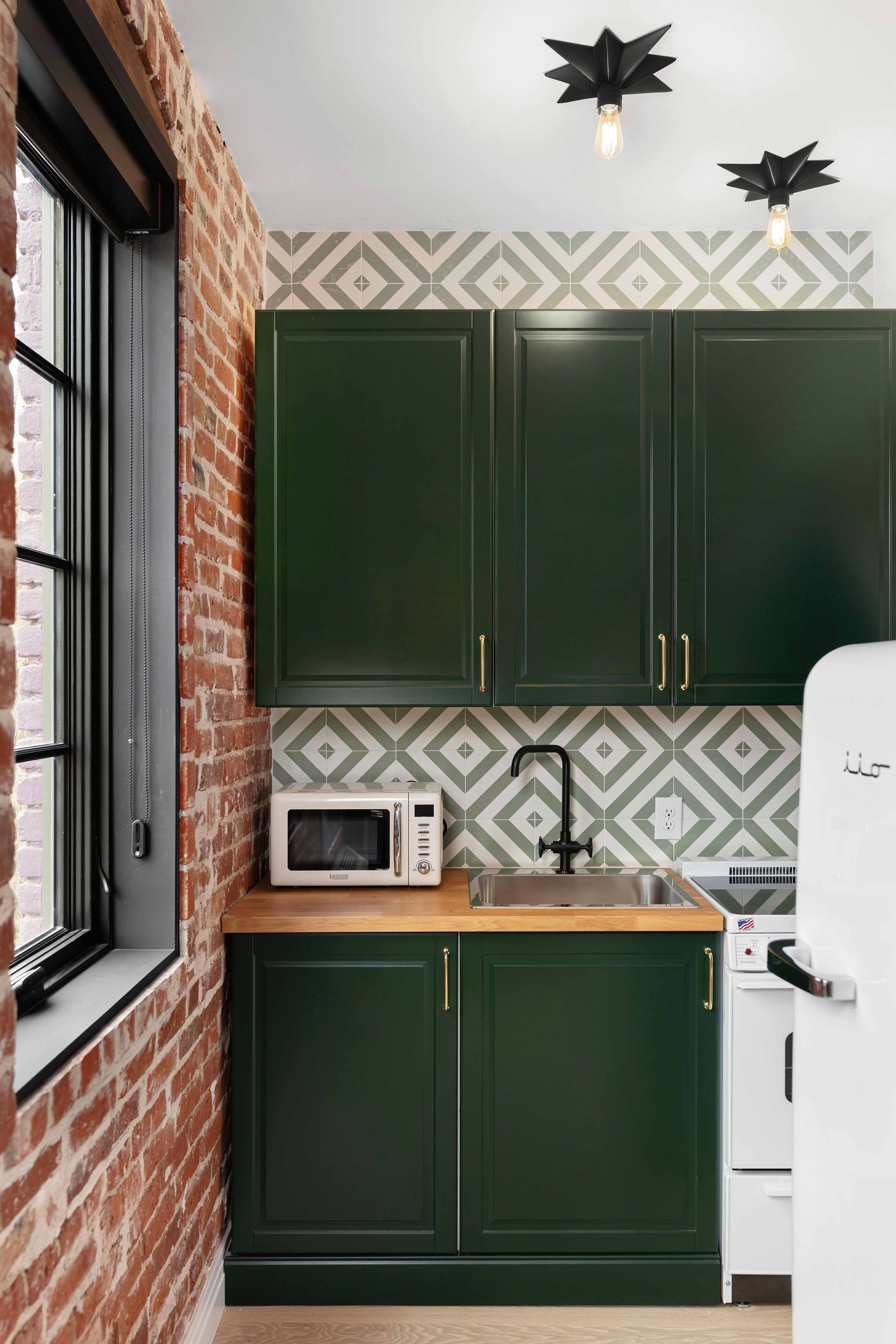 Small kitchen with dark green cabinets, patterned backsplash, exposed brick, and a window on the left.