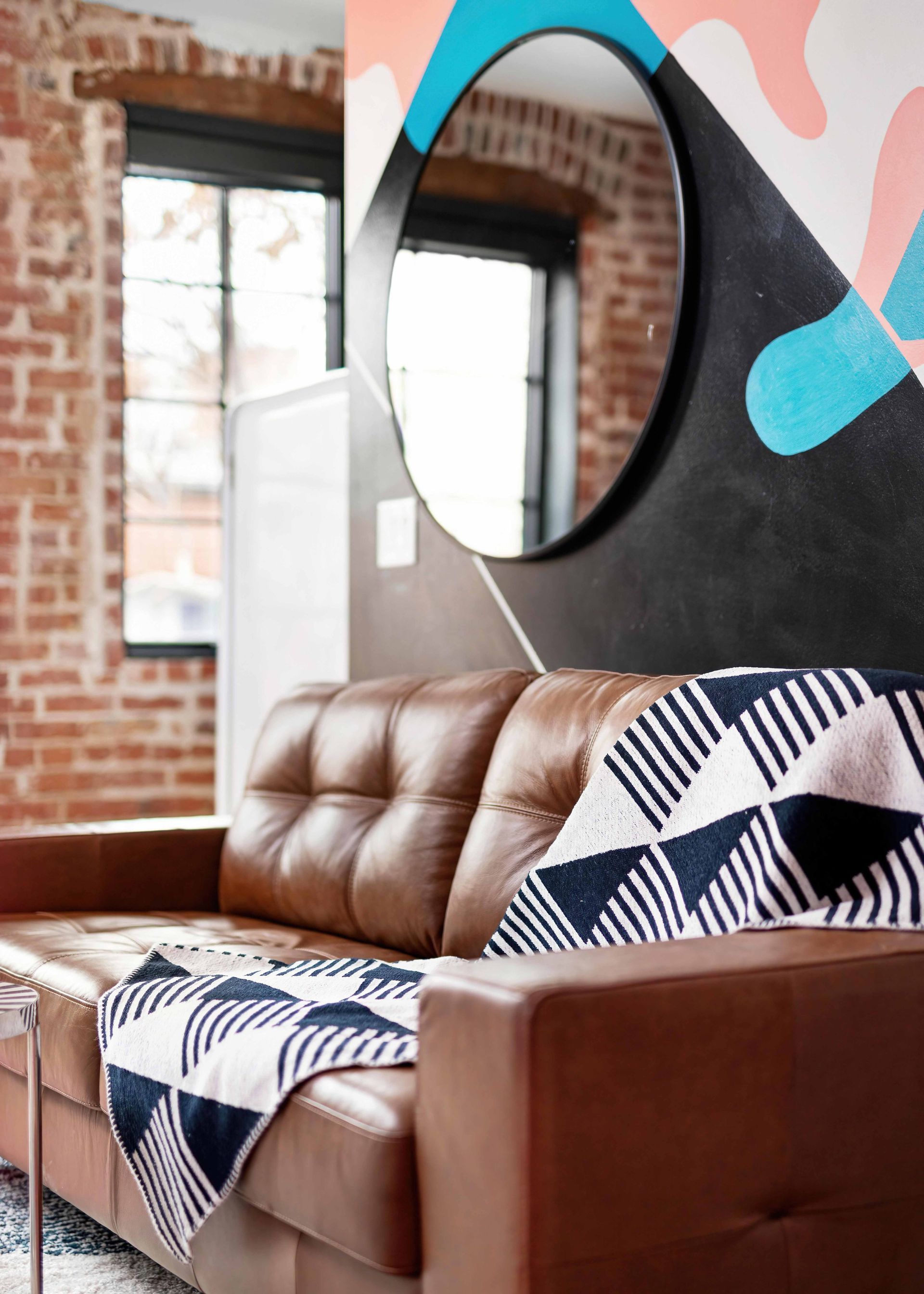 Brown leather sofa with patterned blanket and pillow in a bright room with brick walls and a round mirror