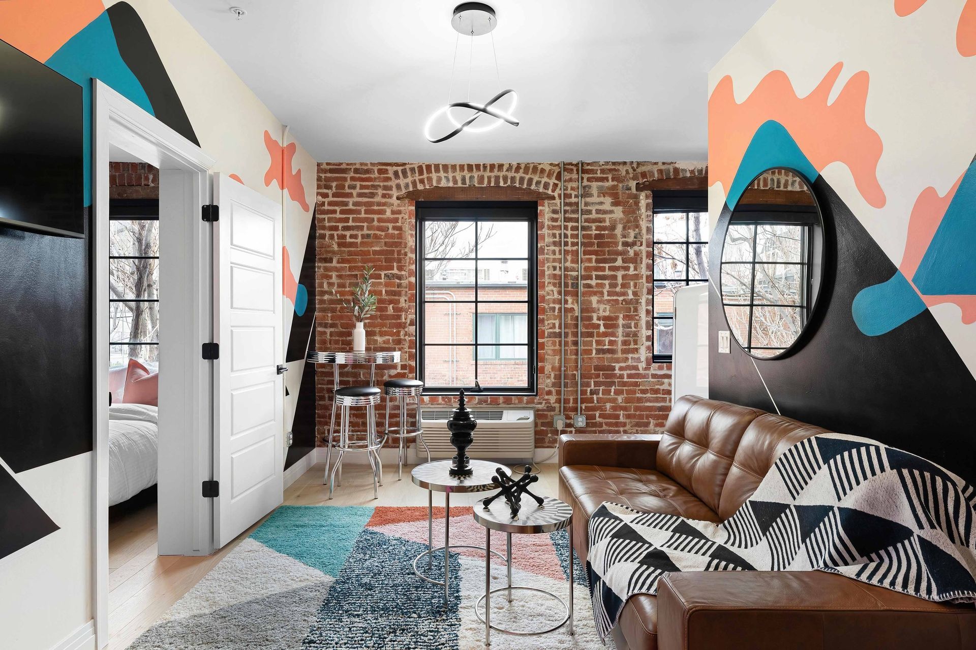 Bright living room with colorful abstract wall art, brown leather sofa, checkered rug, and exposed brick window wall
