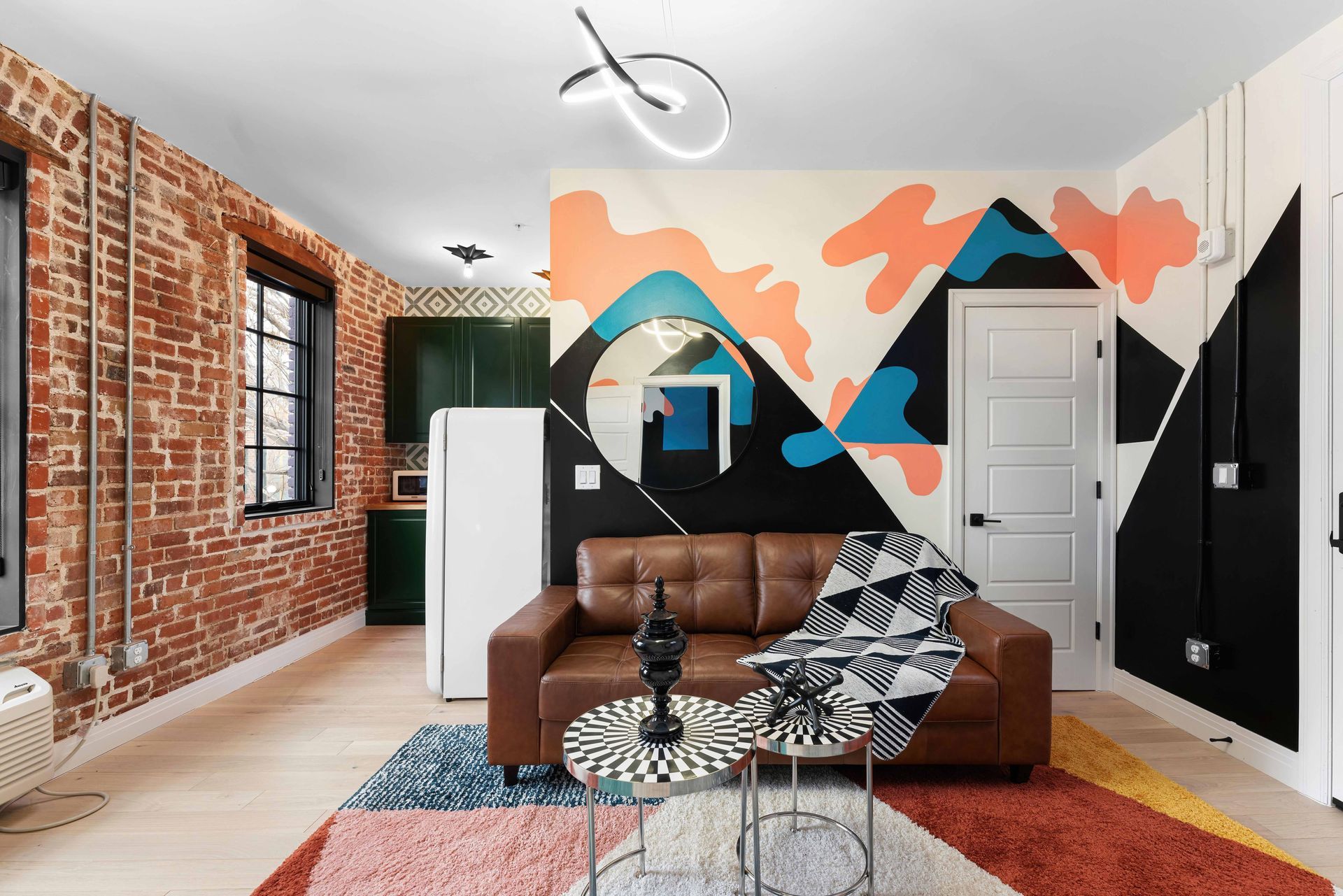 Modern living room with brown sofa, geometric mural, exposed brick wall, and colorful rug
