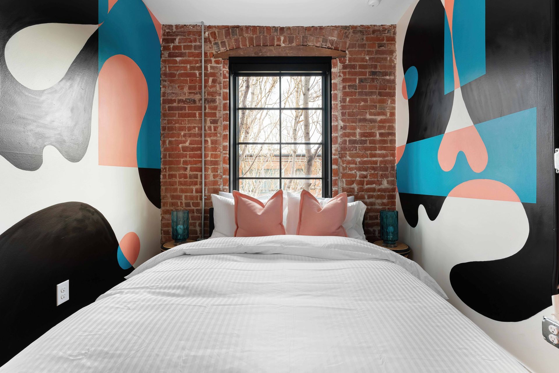 Bedroom with brick accent wall, colorful abstract mural, window, and white bed with pink pillows