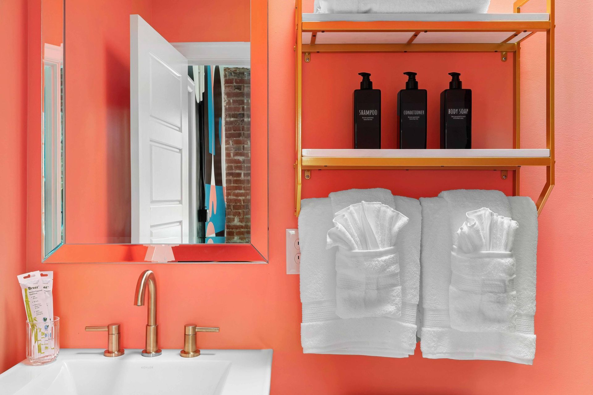 Coral bathroom with mirror, brass faucet, toiletries, black bottles, and towels on shelves