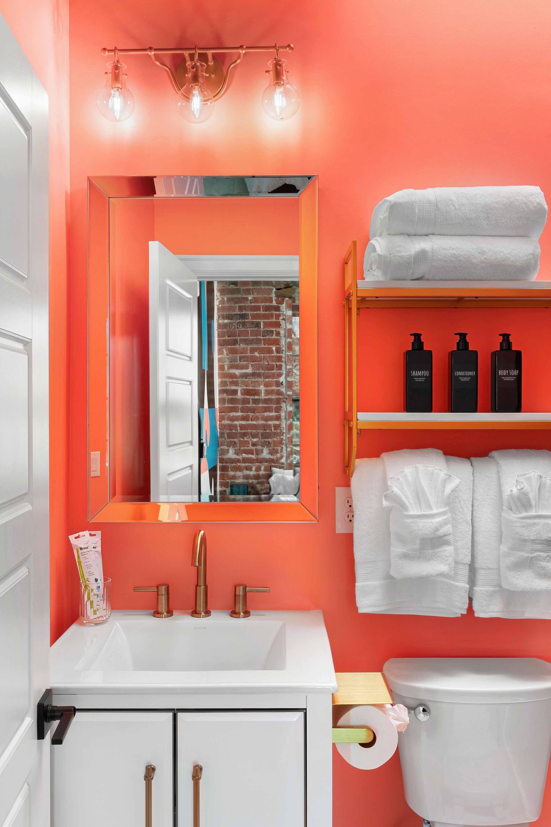 Bright coral bathroom with white sink, toilet, mirror, shelves, towels, and exposed brick doorway