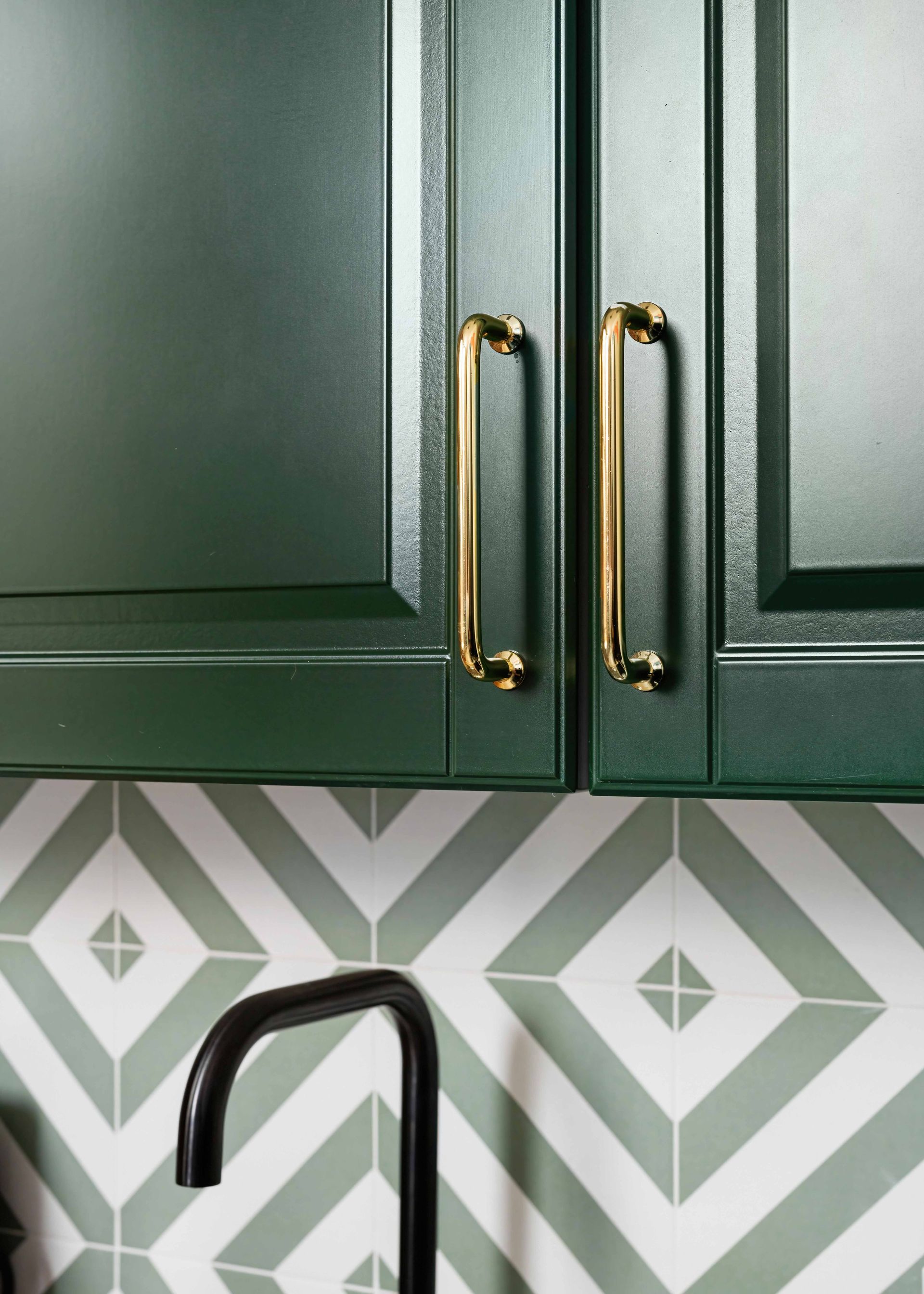 Dark green kitchen cabinets with brass handles above a patterned tile backsplash and black faucet