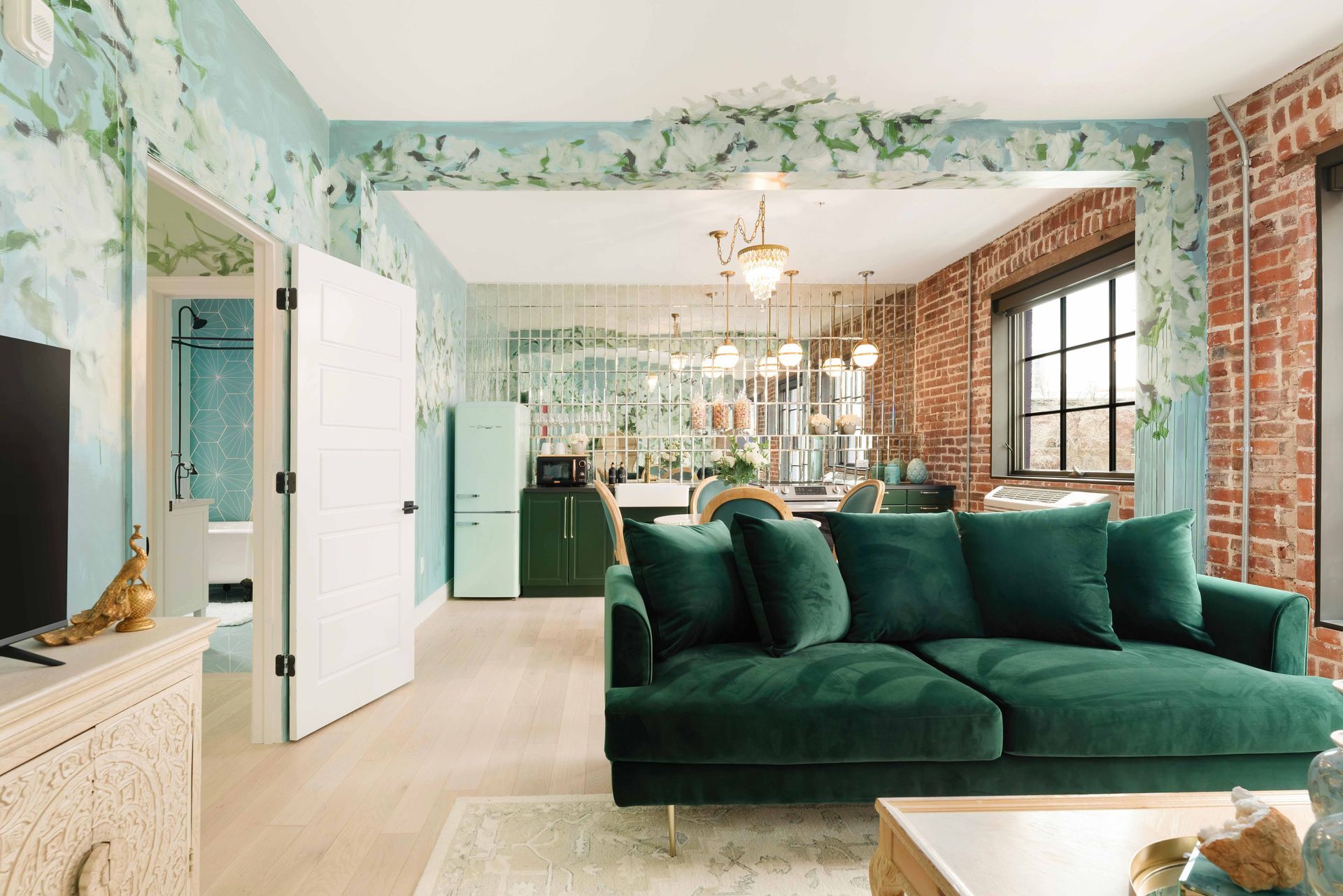 Bright living room with a dark green sofa, floral wallpaper, and open door leading to a kitchen