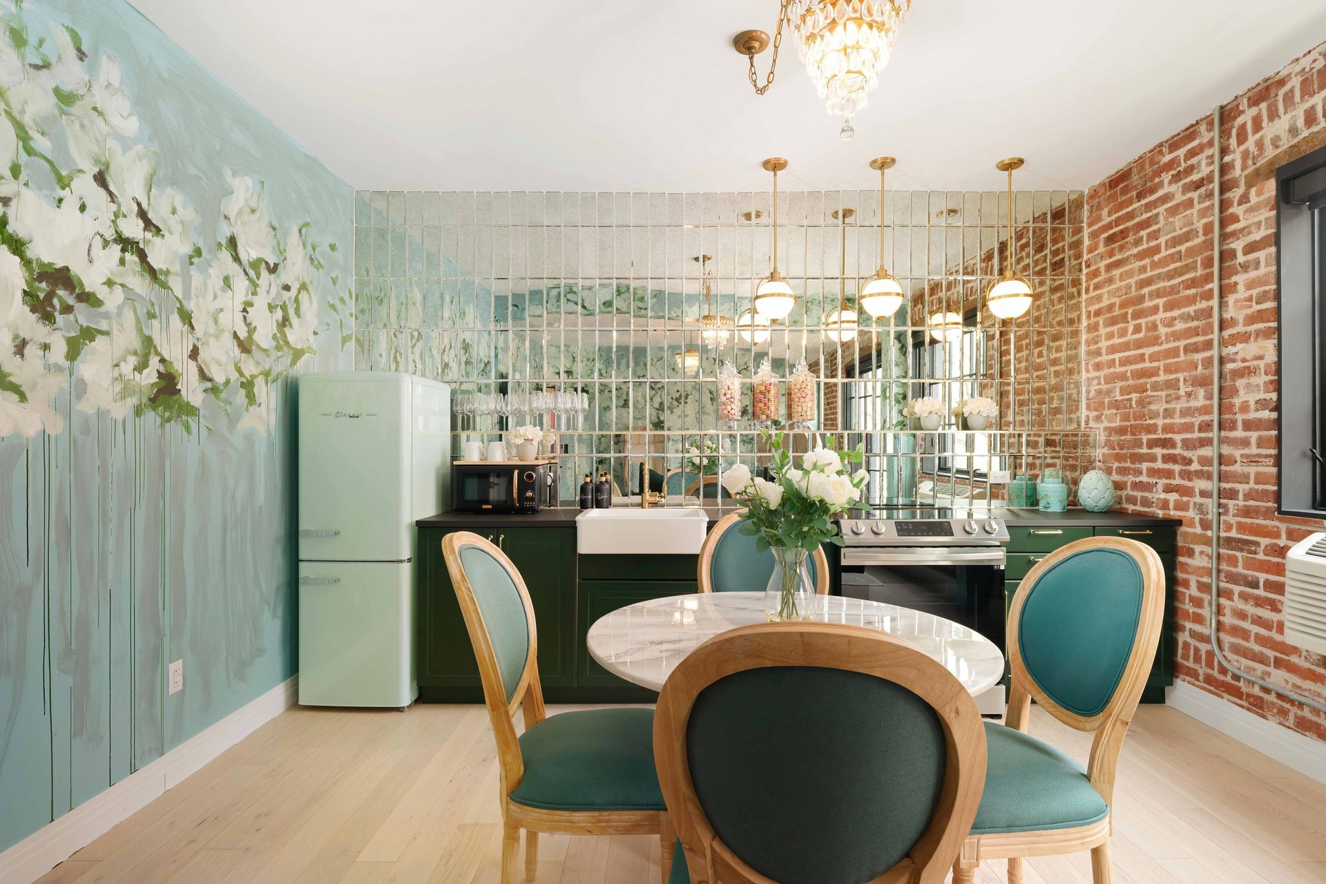 Elegant dining room with teal chairs, round table, brick wall, floral mural, and gold chandeliers