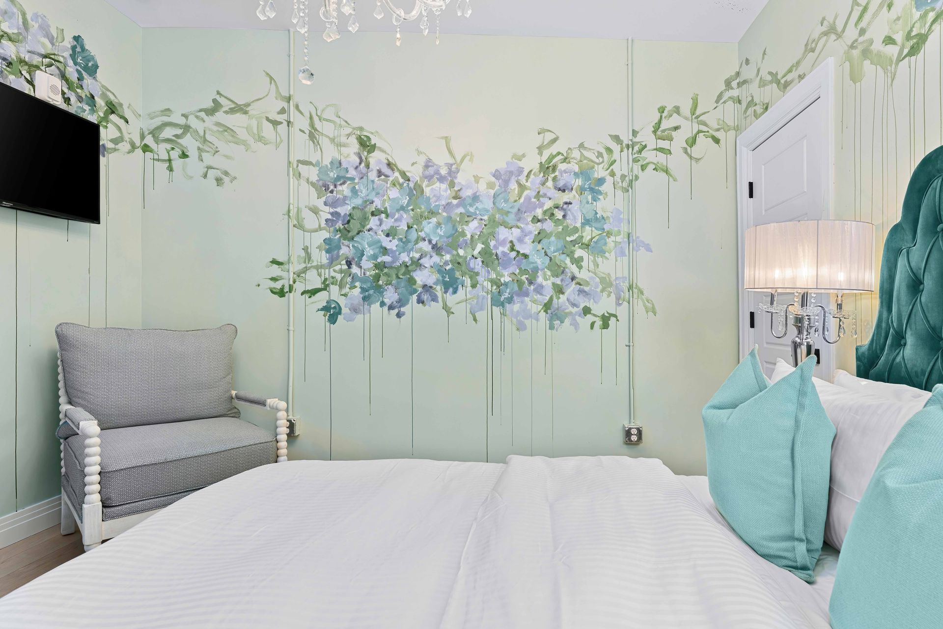Bright bedroom with floral mural, white bed, gray chair, and teal pillows