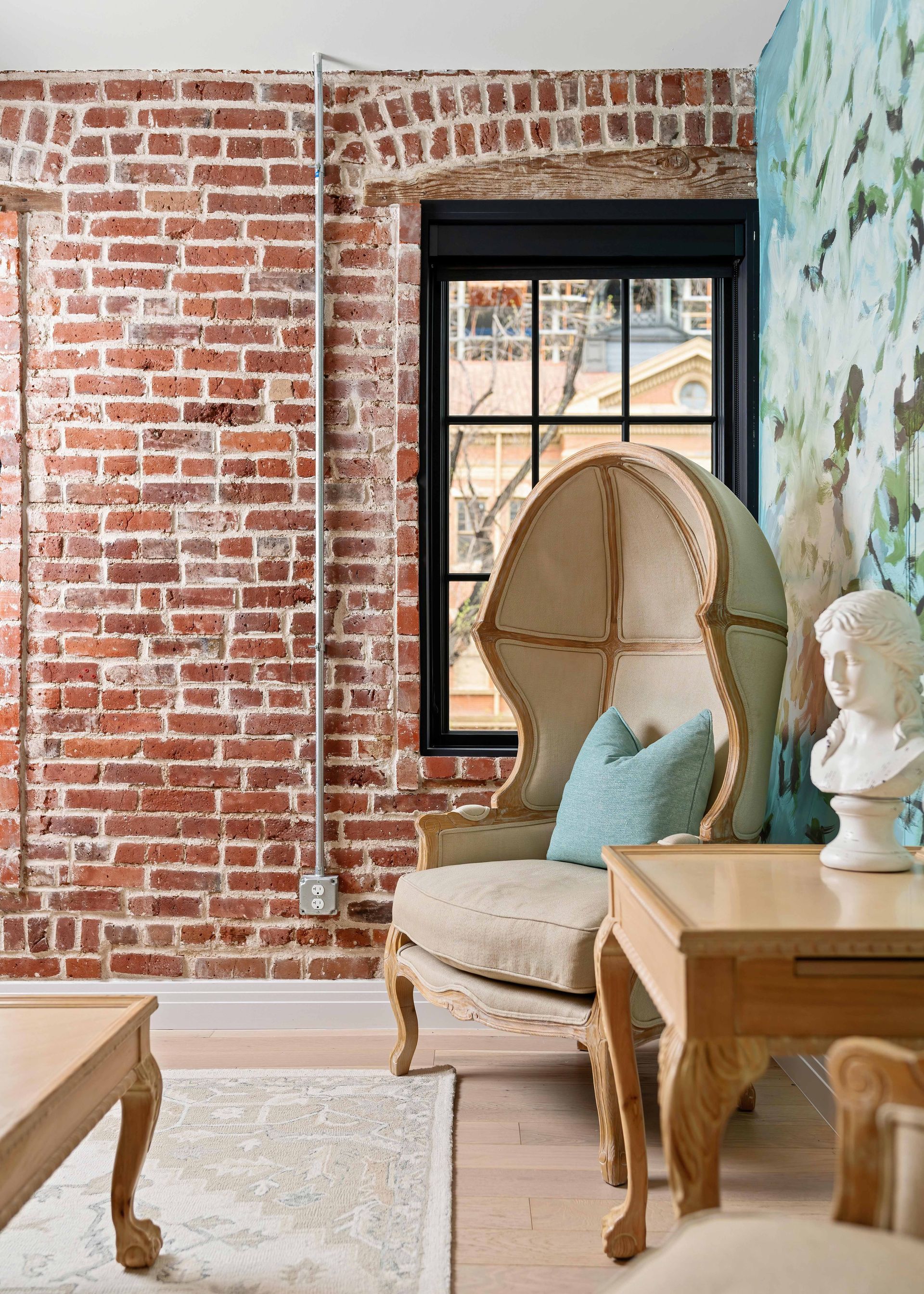 Cozy room with exposed brick wall, black-framed window, cream armchair, and wooden table with bust statue