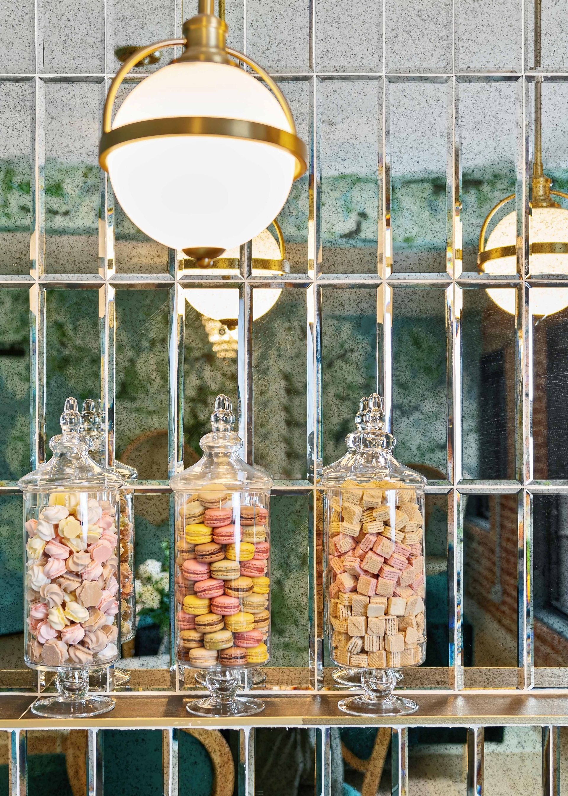 Glass jars of colorful candies on a mirrored shelf beneath round brass pendant lights